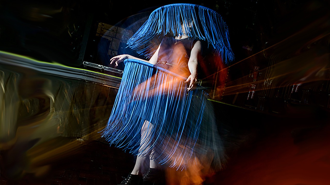 A person stands wearing an avant-garde costume covered in long blue fringe, obscuring their face and upper body, with dramatic lighting and abstract colorful streaks in the background.
