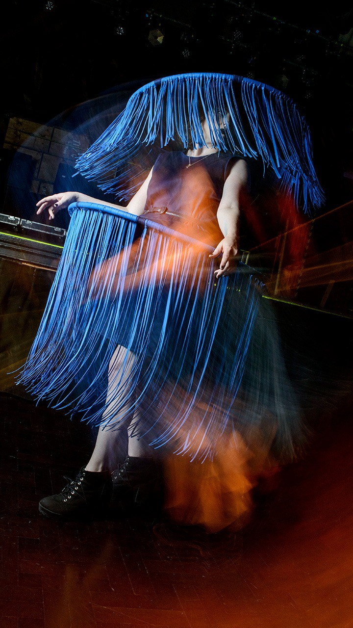 Person wearing a dress and large hat made of long blue fringe, partially obscuring their face and body, creating a dynamic, flowing effect in a dimly lit room.