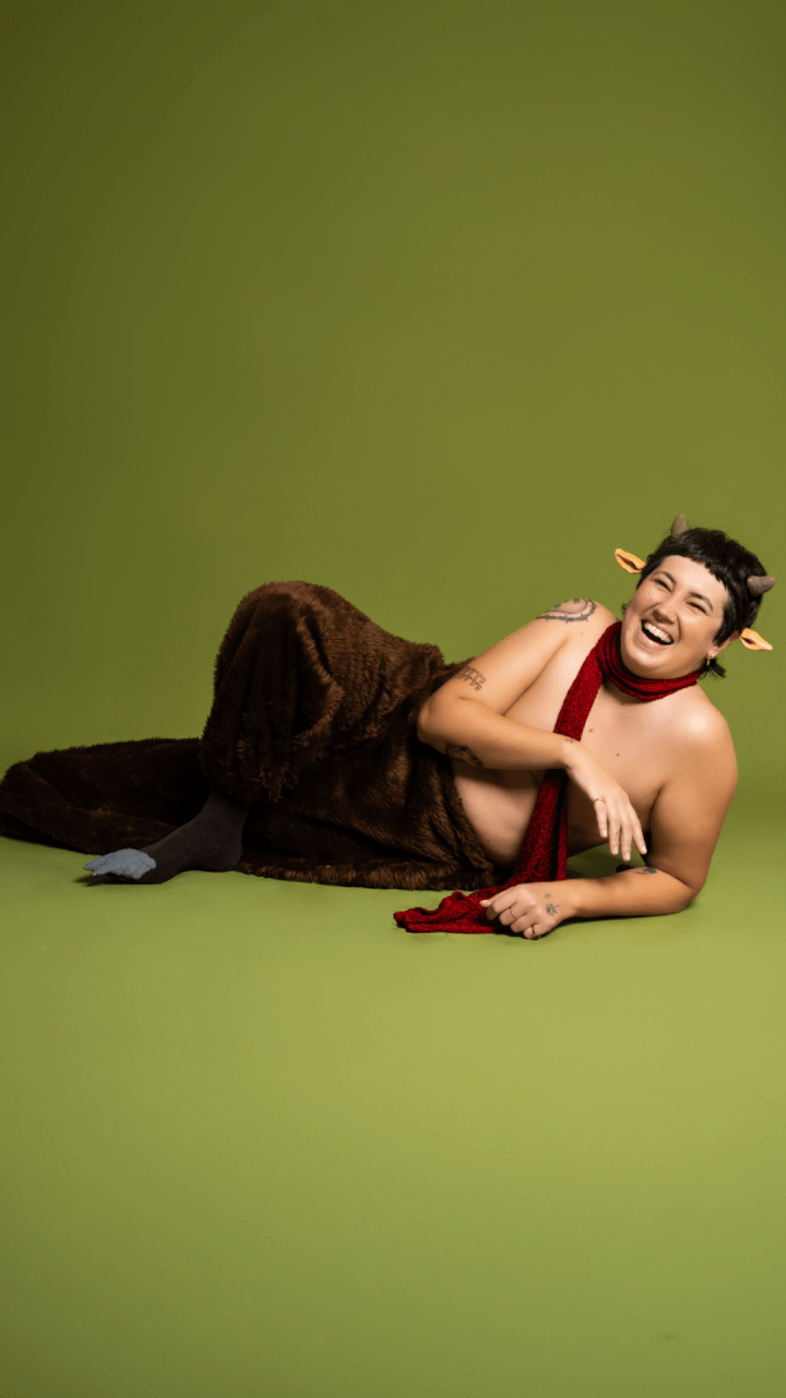 A person dressed as a satyr with furry brown pants, goat ears and horns, and a red scarf, lies on their side against a green background, smiling and laughing.