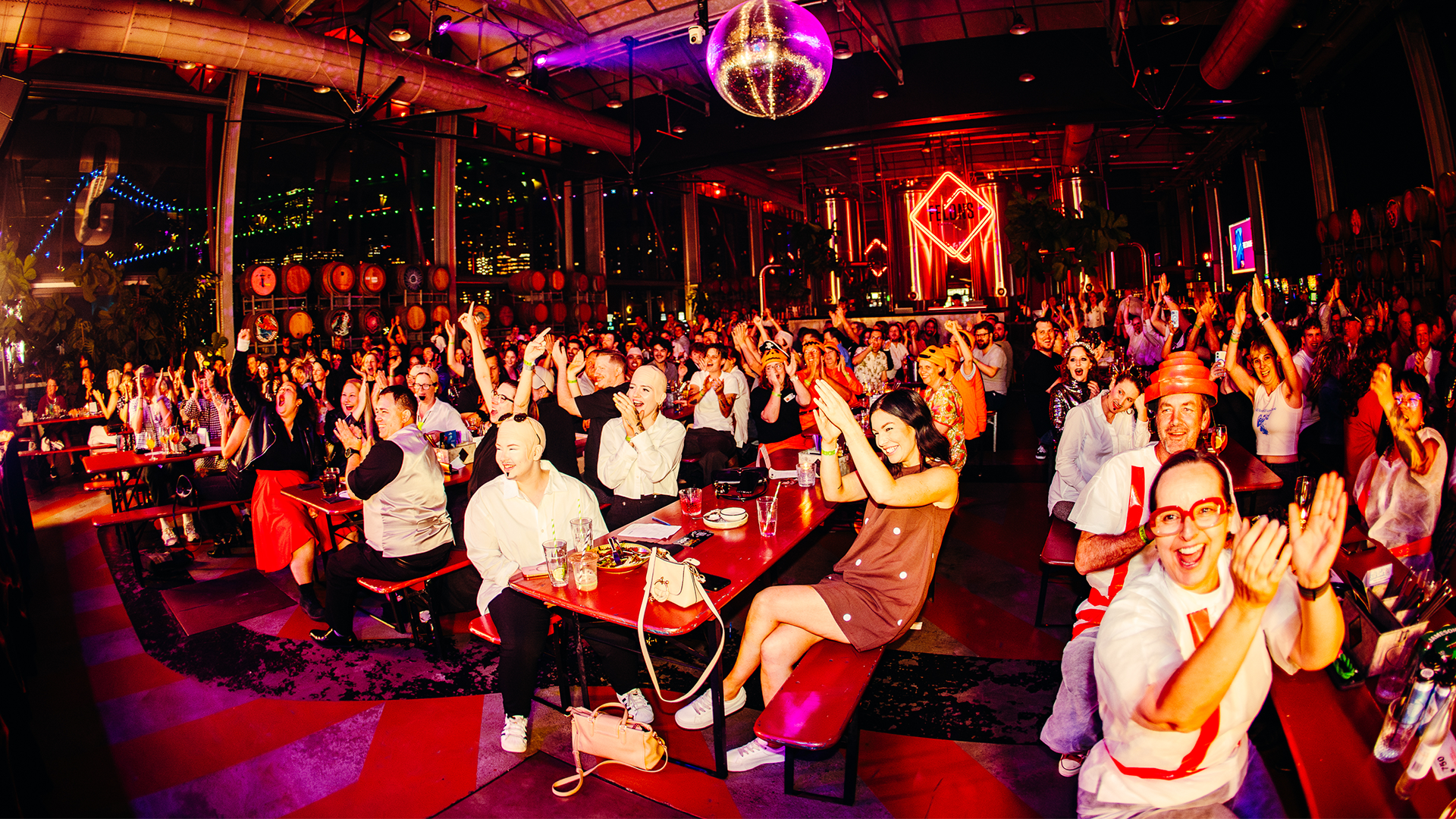 A lively crowd sits at tables in a vibrant, neon-lit venue, cheering and clapping with excitement. A disco ball hangs above, and people smile and enjoy drinks, creating a festive party atmosphere.