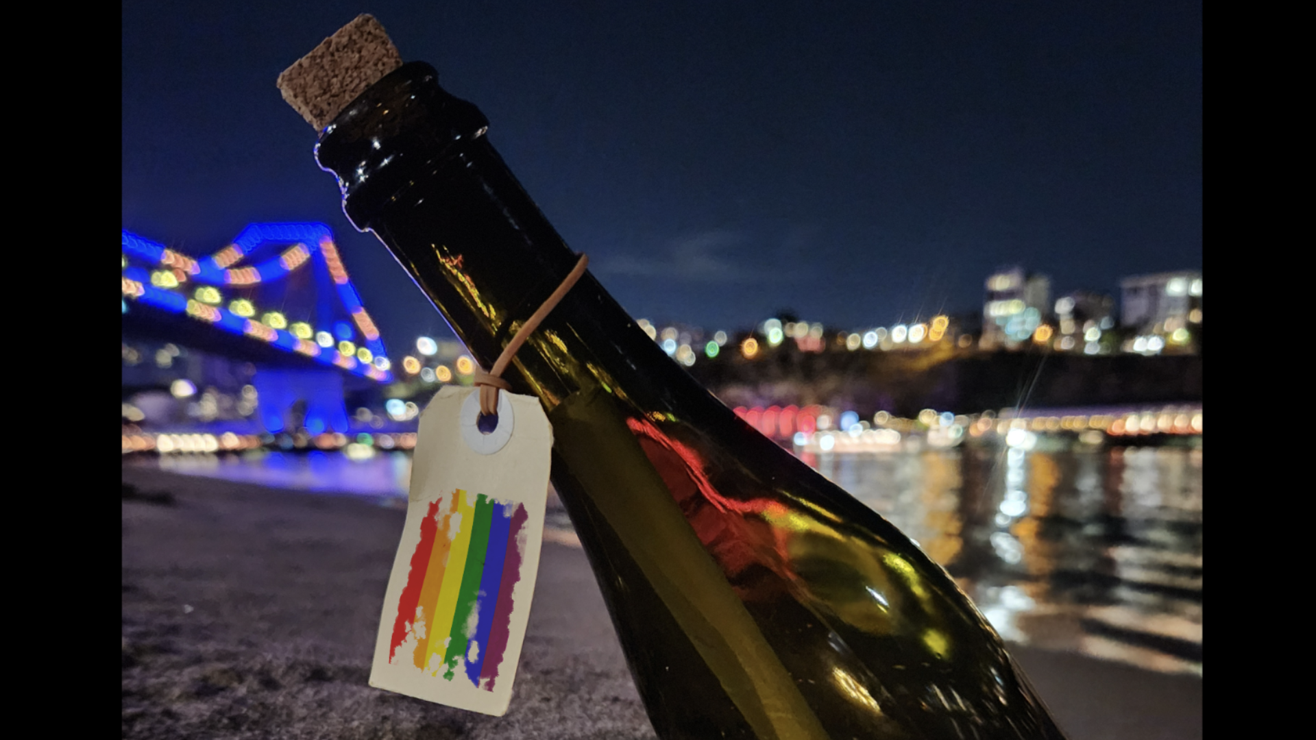 A corked wine bottle with a rainbow tag lies on sand near a river at night. City lights and a lit-up bridge are blurred in the background, reflecting on the water.