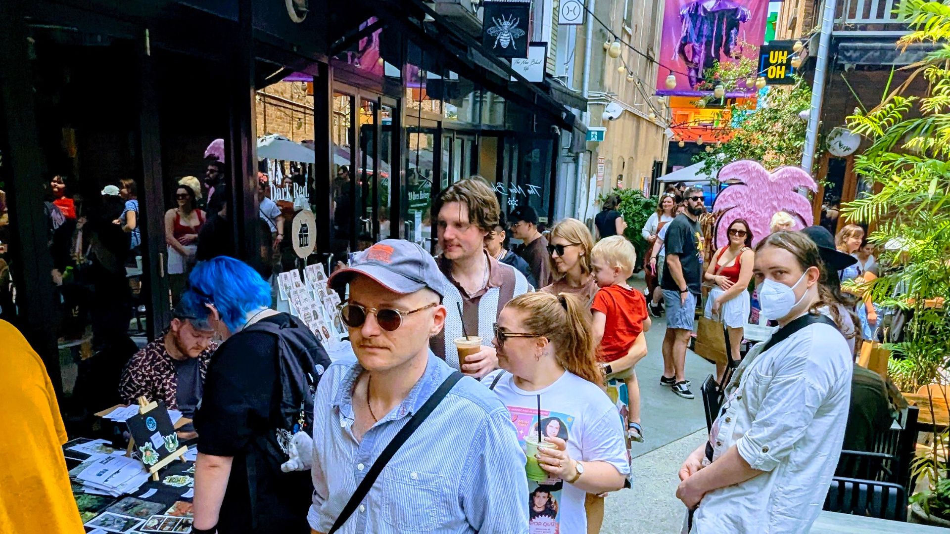 A busy street scene shows people walking past shops and outdoor tables. Some hold drinks, one person wears a mask, and colorful banners and plants decorate the lively, sunlit area.