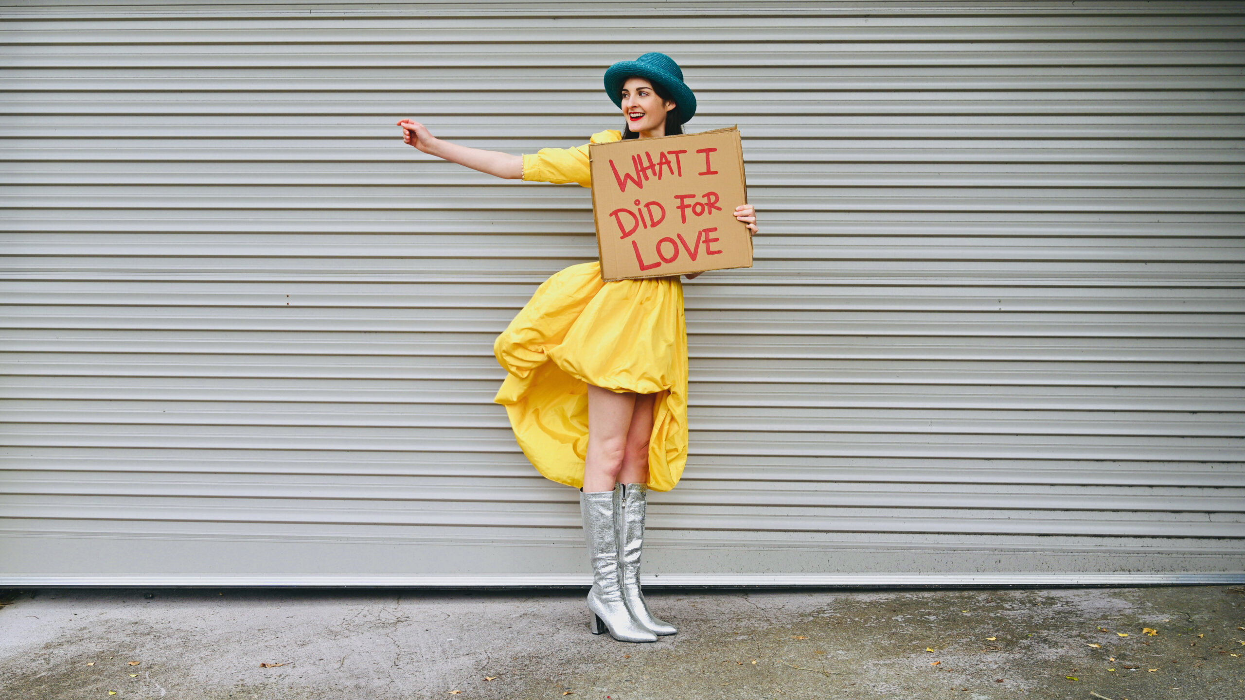 A person in a yellow dress, teal hat, and silver boots holds a cardboard sign that reads “WHAT I DID FOR LOVE” while standing in front of a gray metal garage door.