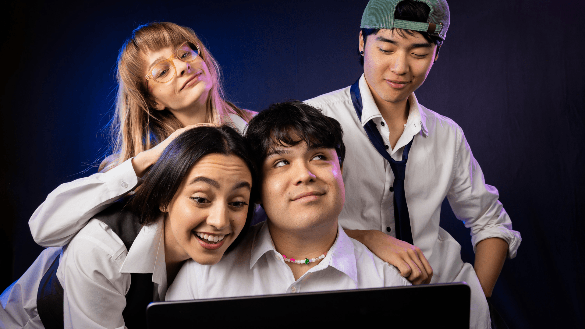Four young adults in white shirts gather closely around a laptop, smiling and laughing. One wears glasses, another a backwards cap, and the group appears cheerful and engaged, with a dark background behind them.