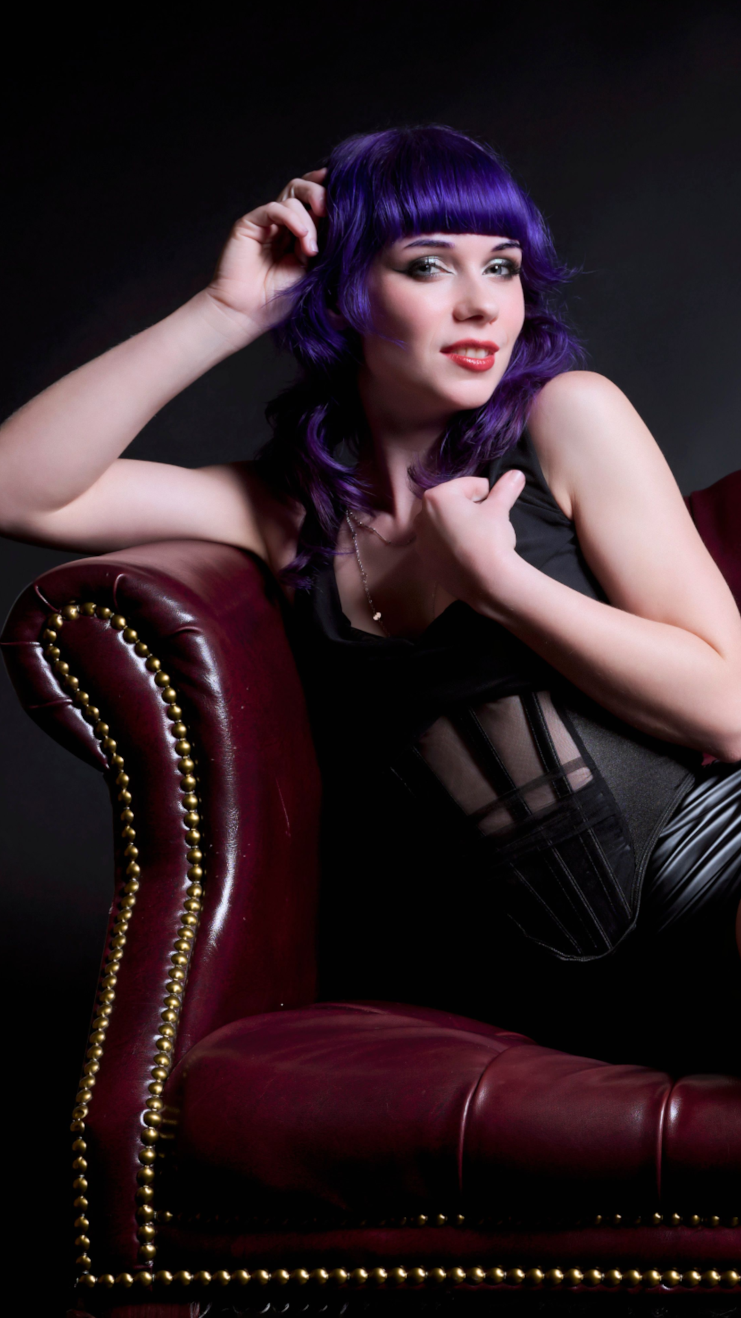 Woman with wavy purple hair and red lipstick poses on a dark red leather armchair, wearing a black outfit with sheer panels, looking at the camera with one hand on her head and the other on her chest, set against a dark background.