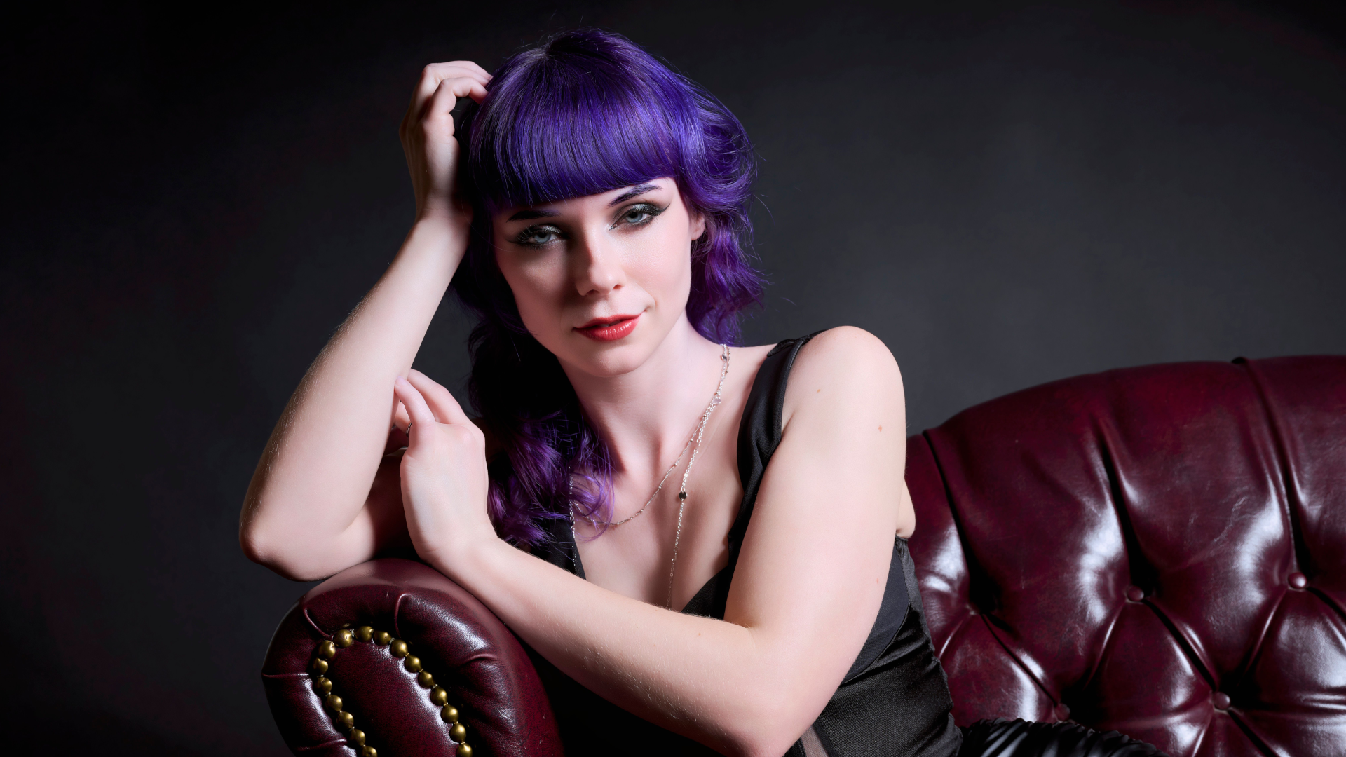 A woman with purple hair and bangs sits on a maroon leather chair, resting her head on one hand. She wears a black sleeveless top and gazes directly at the camera against a dark background.