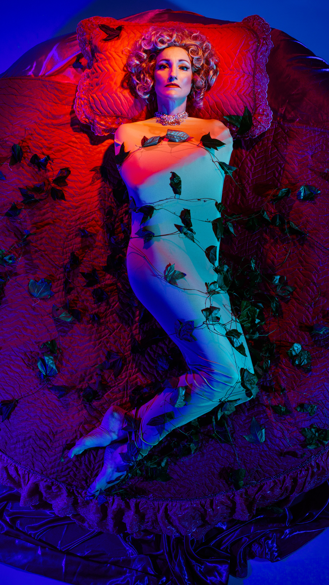 A woman in a glamorous gown lies on a red quilted bed, wrapped in ivy vines, with dramatic red and blue lighting and vintage-style hair and jewelry.