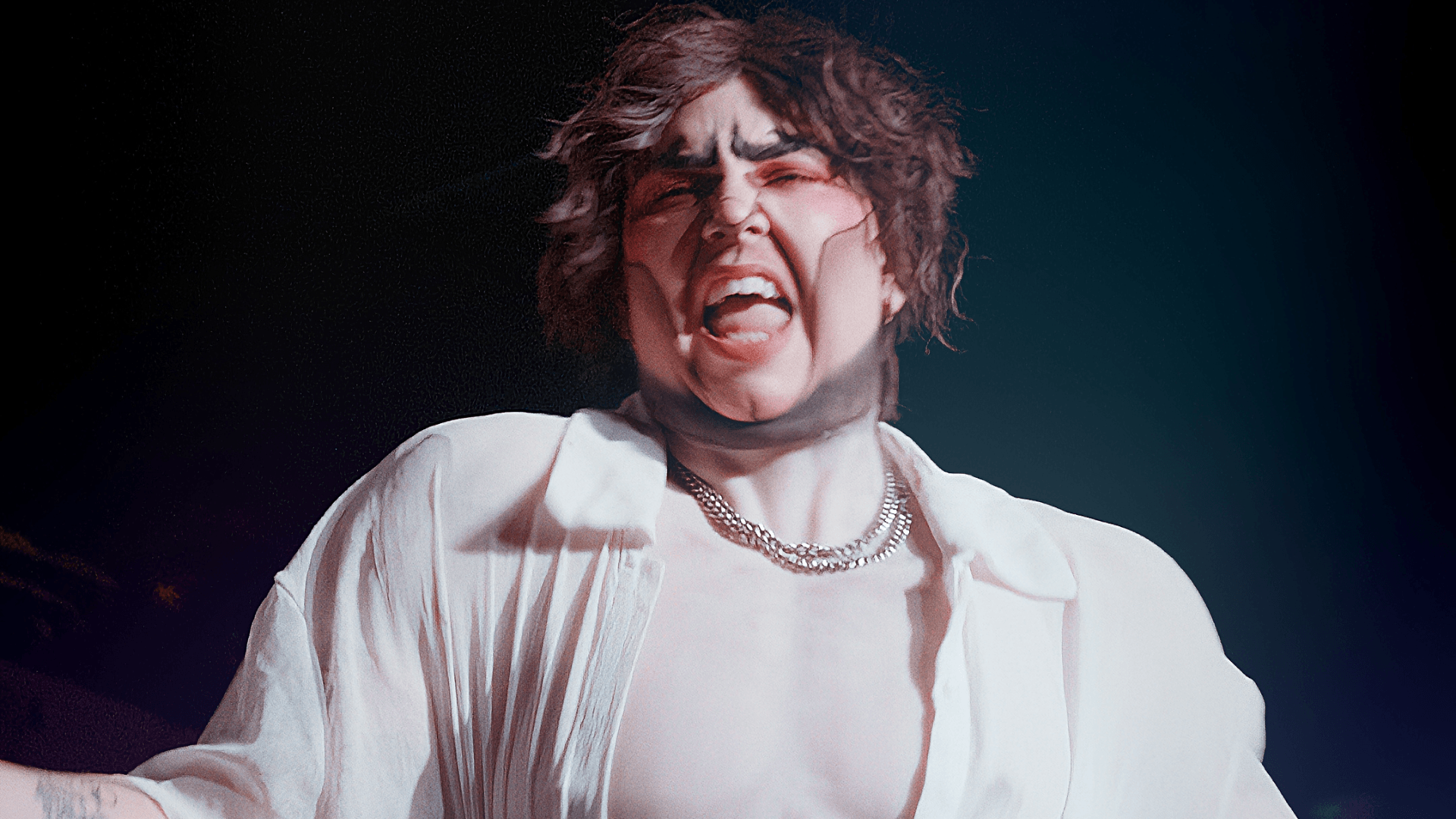 Person with short, dark, tousled hair wearing a white shirt open at the chest and a chain necklace, singing or shouting expressively on a dimly lit stage.