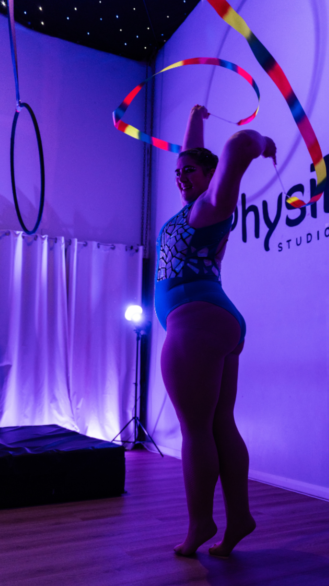 A performer in a blue leotard poses with a colorful hoop under purple stage lighting at a studio, with an aerial ring and curtains in the background.