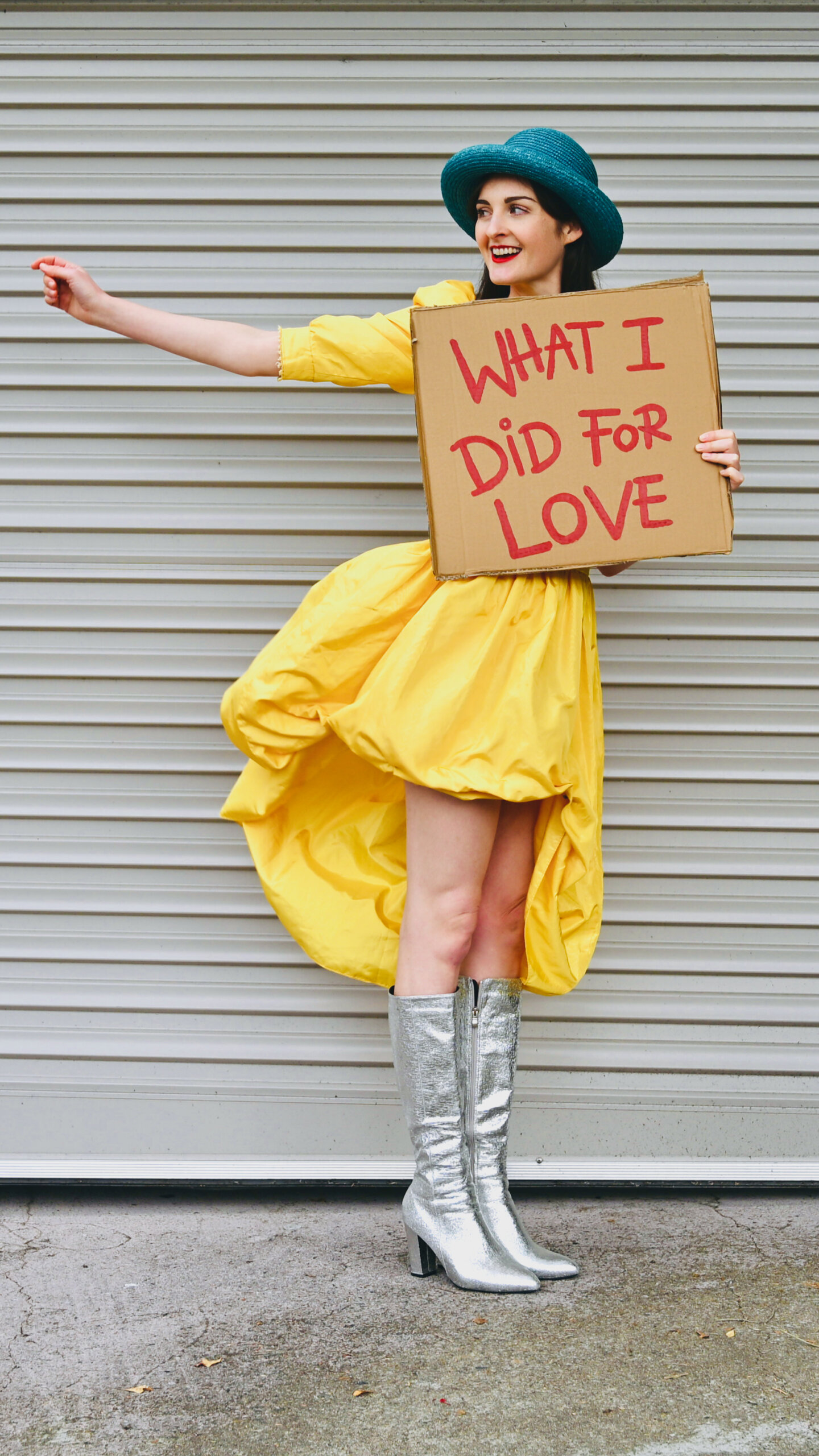 A person in a yellow dress, teal hat, and silver boots holds a cardboard sign reading WHAT I DID FOR LOVE while smiling and posing in front of a gray metal door.