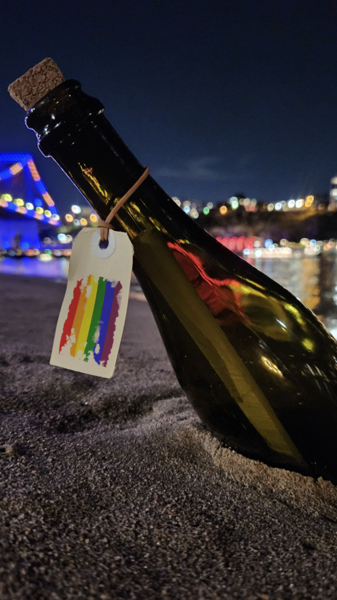 A wine bottle with a rainbow tag is partly buried in sand at night, with city lights and a brightly lit bridge in the background, reflecting on water.
