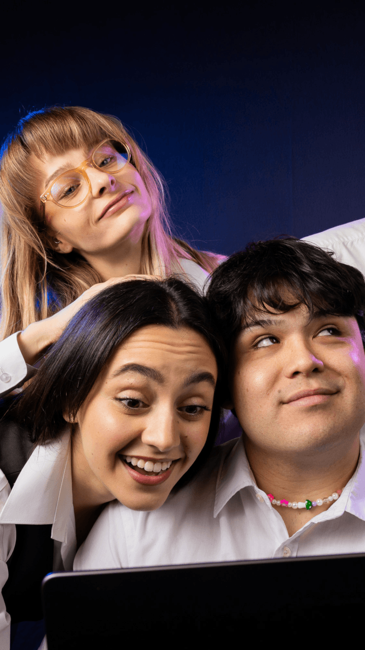 Three young adults, two women and one man, smiling and posing together in front of a dark background. They are wearing white shirts and appear happy and engaged, with a laptop visible in the foreground.
