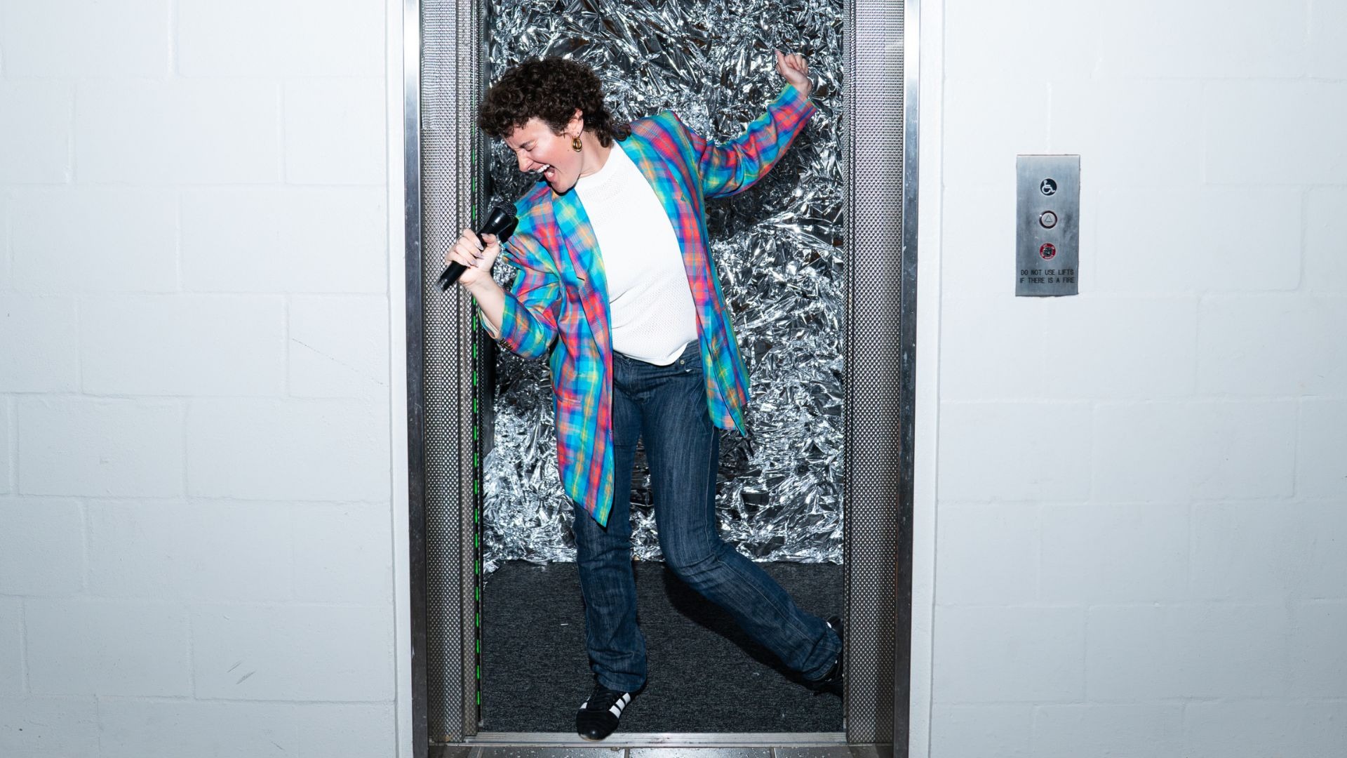 A person in a colorful jacket sings into a microphone while standing in an open elevator with a shiny, foil-like background.