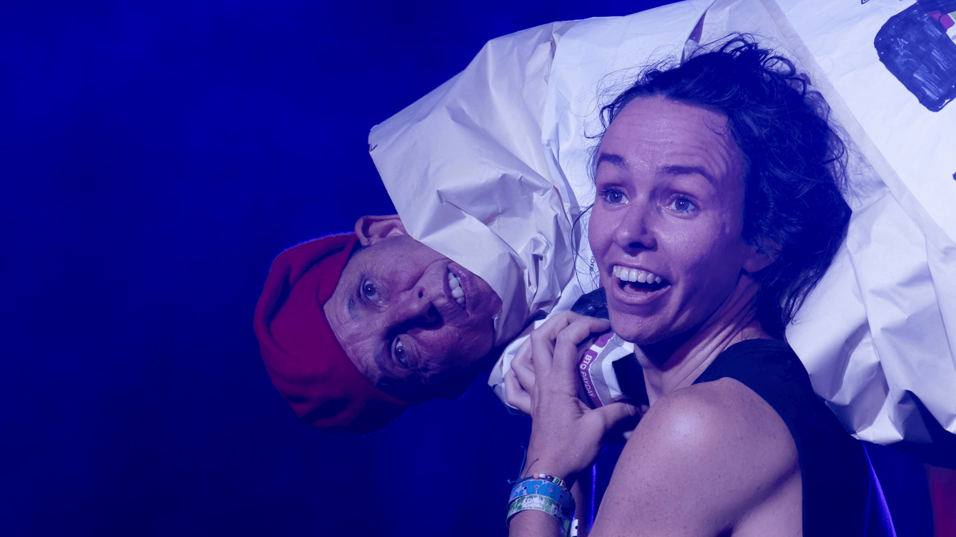 A woman smiling holds a person in a red beret draped in white fabric over her shoulder, both appear on stage under blue lighting.