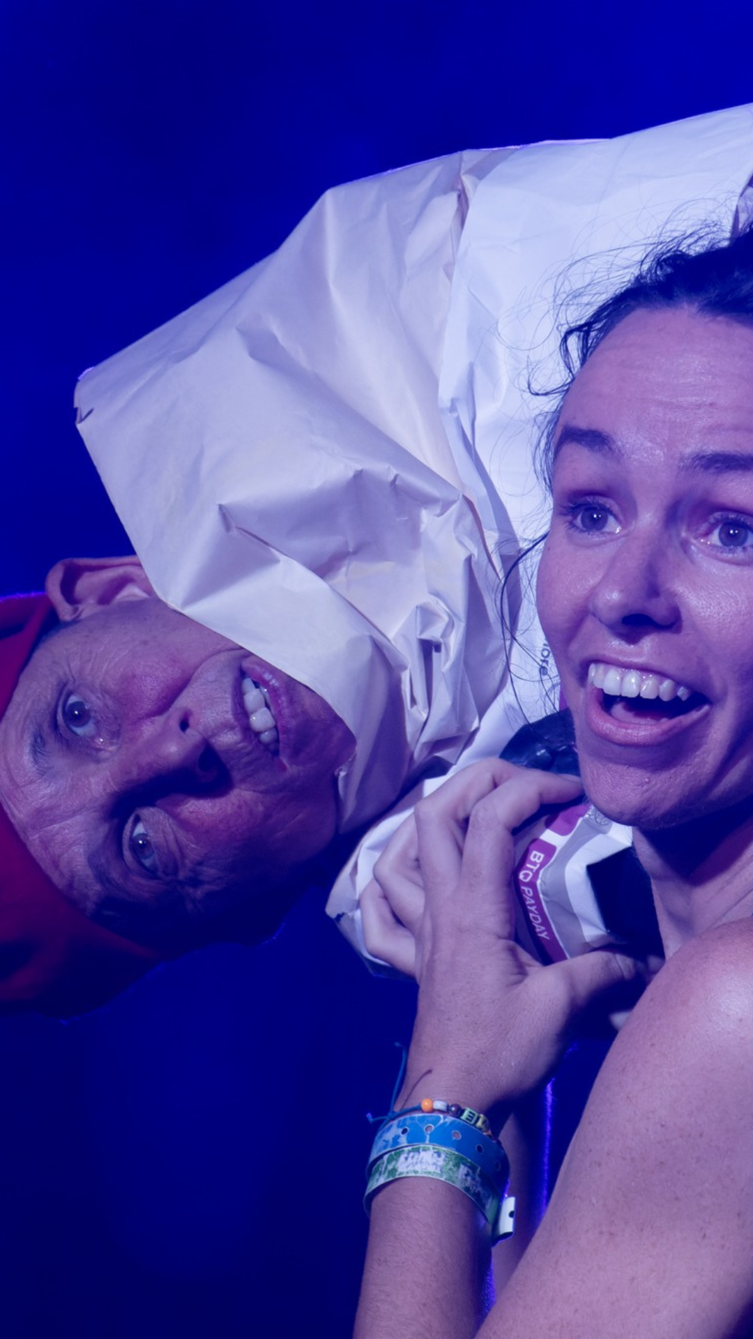 Two people under dramatic blue lighting, one wearing a red hat with their head partially wrapped in crumpled white paper, and the other smiling widely, holding the paper.
