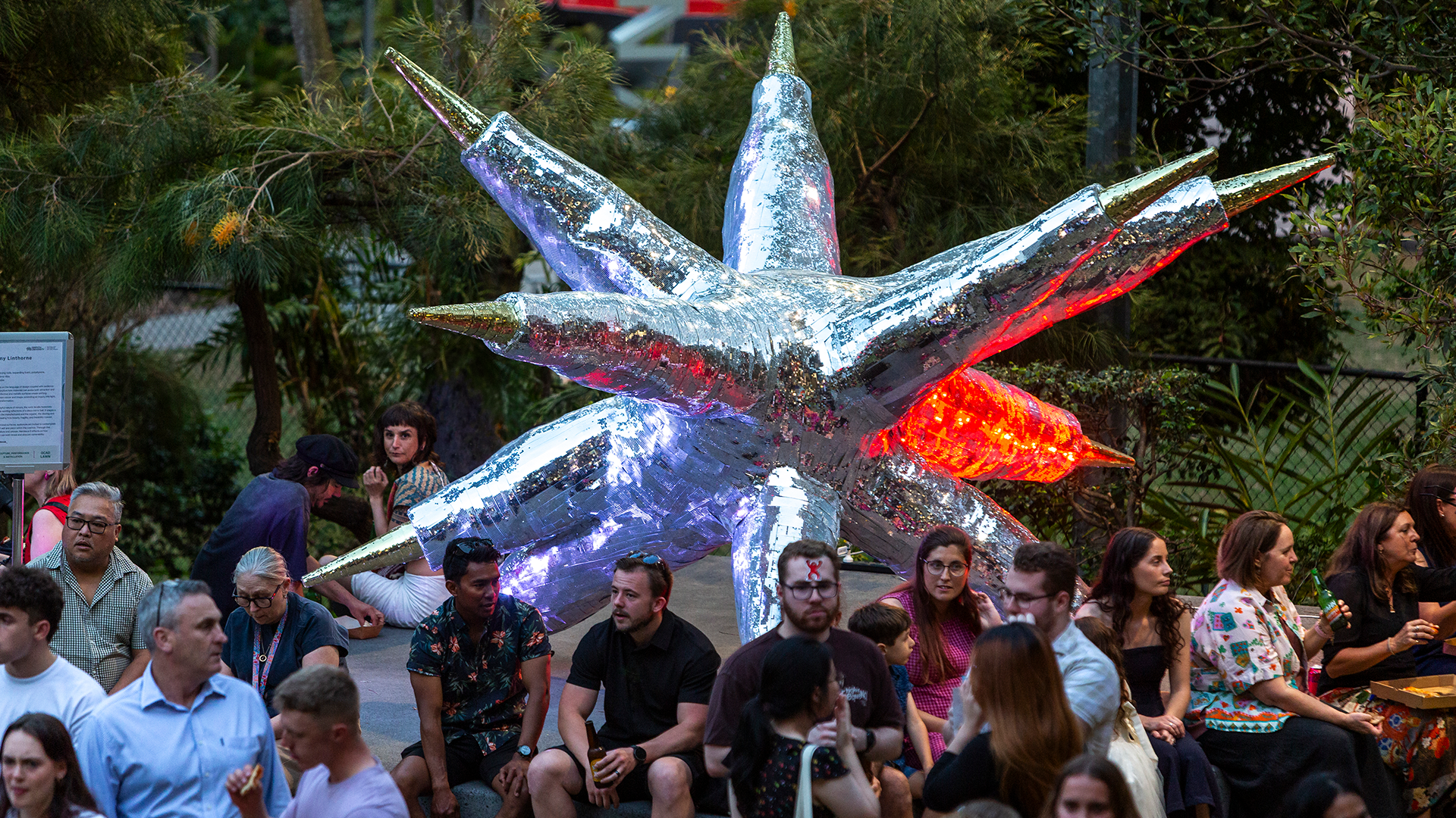 A crowd sits and stands around a large, shiny silver star-shaped sculpture with glowing lights. Trees and plants surround the outdoor area, giving a lively atmosphere.