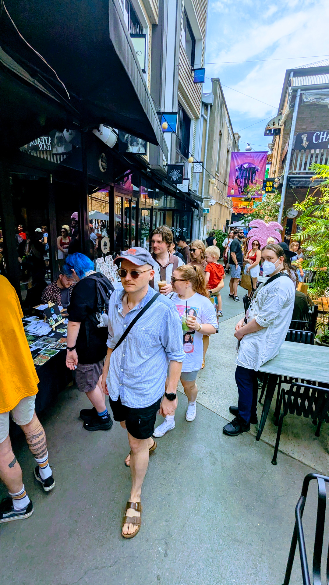 Crowd of people walking down a lively, colorful city street lined with shops and outdoor tables. Some are browsing, others chatting or walking. Bright banners and signs hang above.
