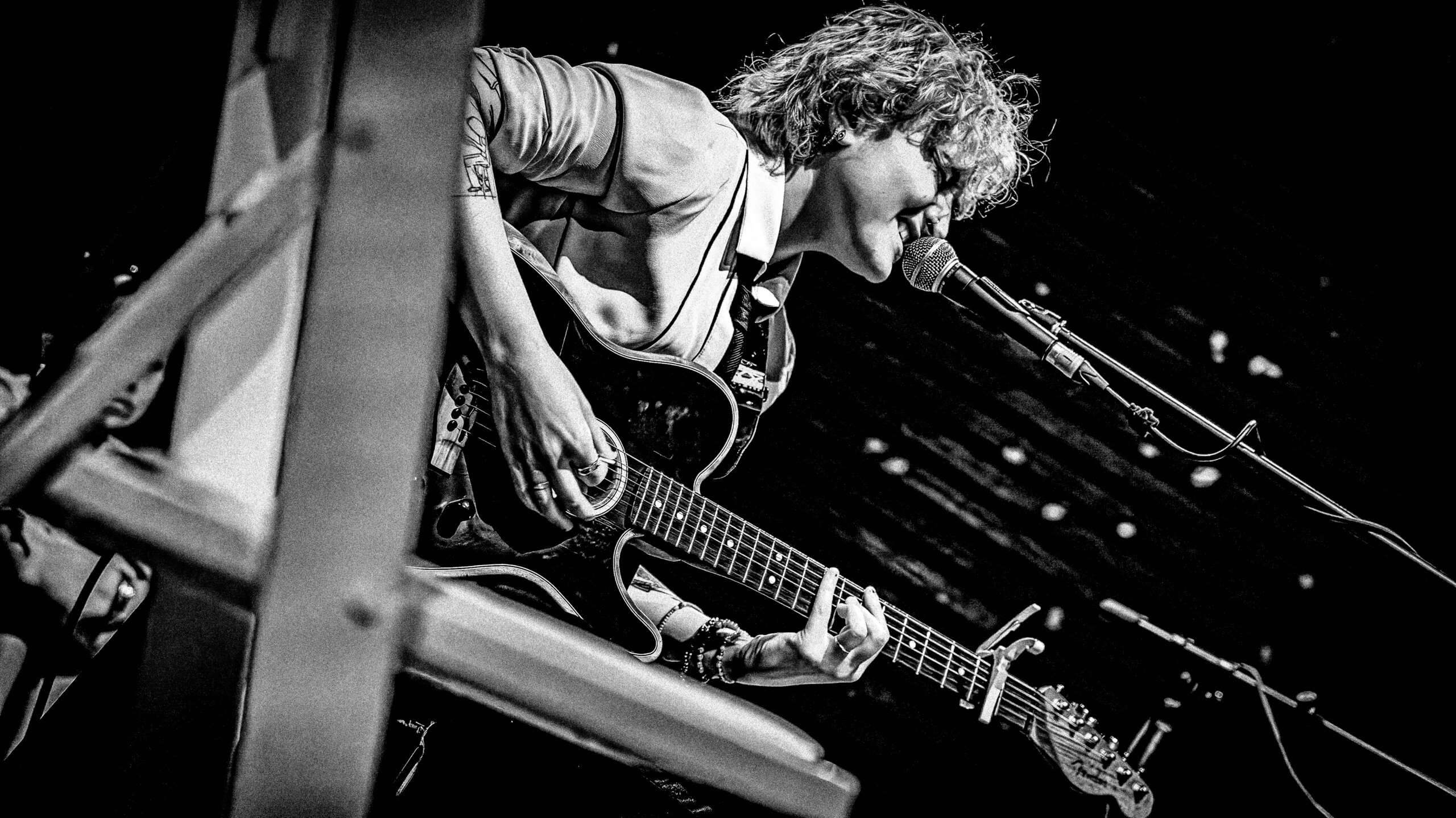 A person with curly hair plays an acoustic guitar and sings into a microphone on stage. The image is in black and white and shot from a low, side angle.