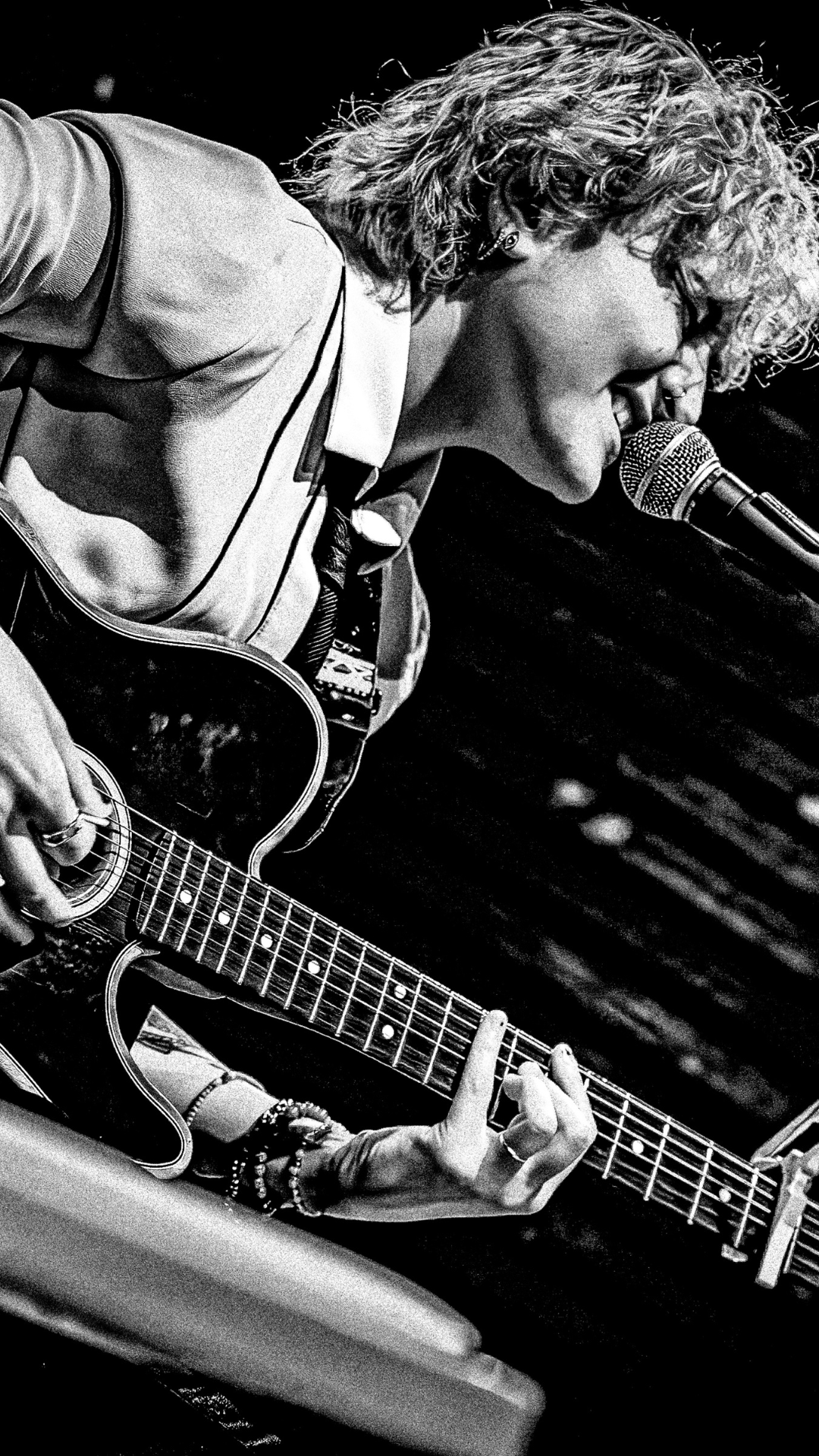Black and white photo of a person with curly hair playing an acoustic guitar and singing into a microphone, wearing a suit jacket and bracelets, performing on stage.