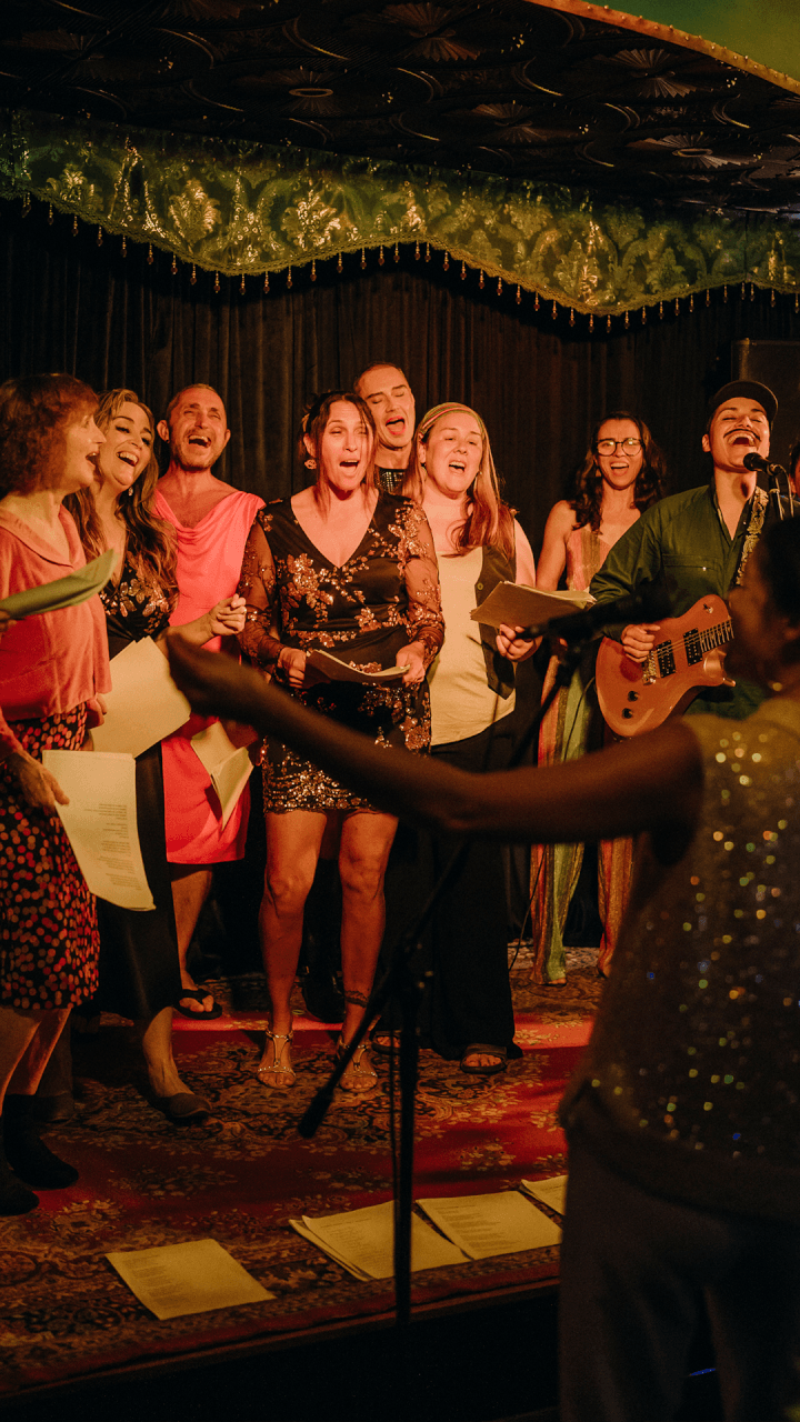 A group of people stand together on a stage, singing enthusiastically with sheets of paper in hand. One person plays an electric guitar while another, with arms raised, faces the group. The scene is warmly lit and lively.