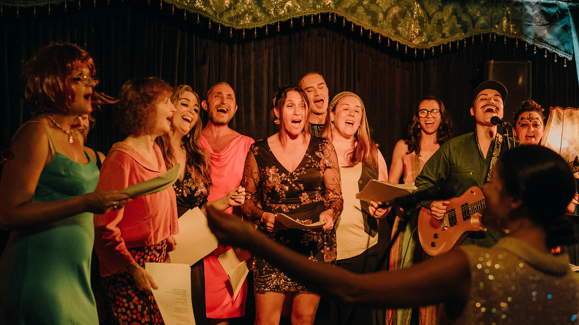 A group of people enthusiastically sing together on stage, some holding papers. They are dressed in colorful, festive clothing, and a person with a guitar stands among them. The background features dark curtains and warm, decorative lighting.