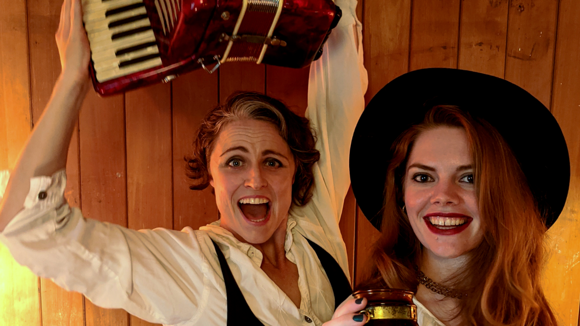 Two smiling people pose in front of a wooden wall. One raises an accordion above their head, while the other, wearing a wide-brimmed hat, holds a drink. Both appear joyful and energetic.