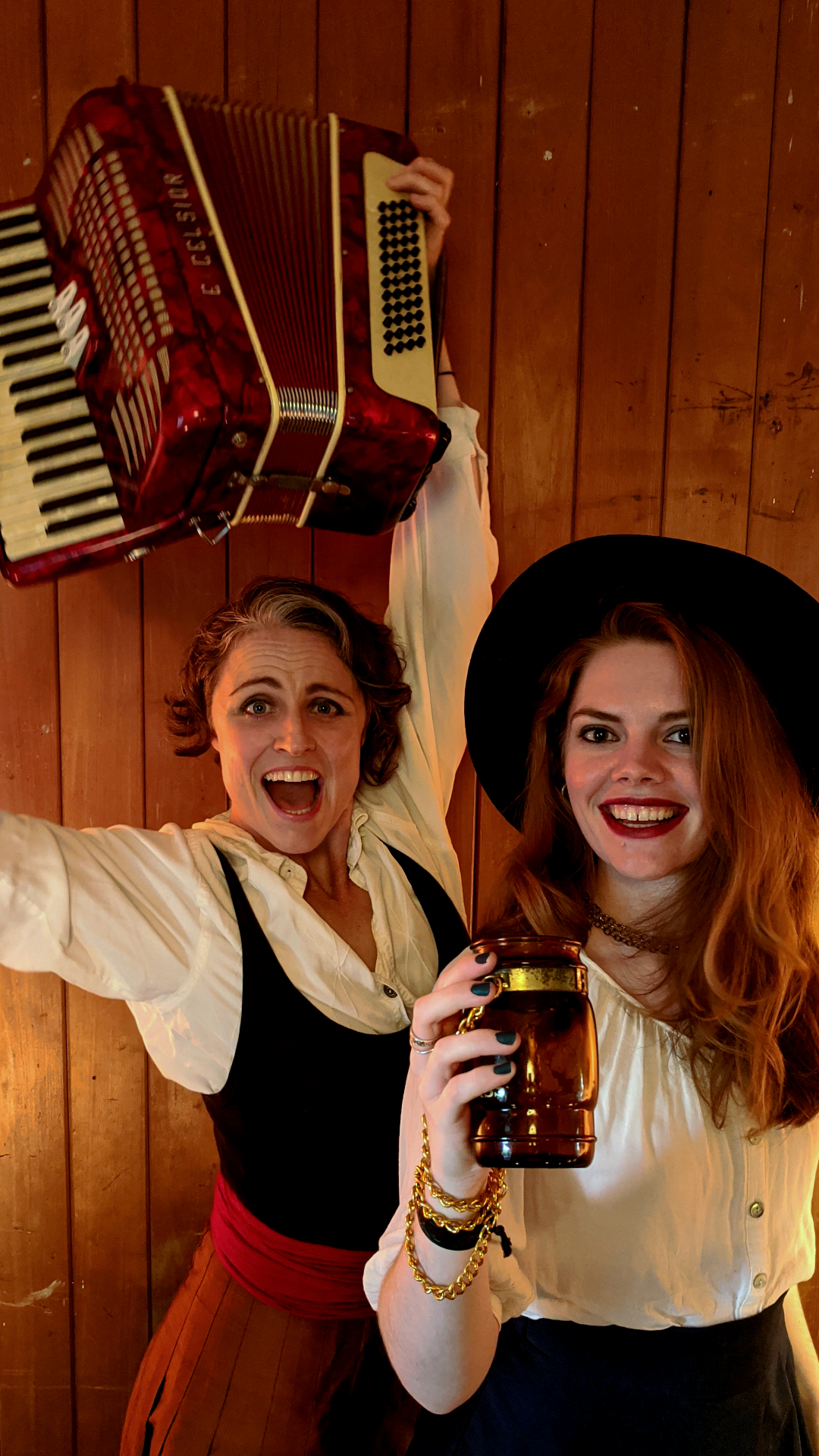 Two women in vintage-style outfits pose joyfully. One holds an accordion above her head, and the other holds a brown jar, smiling. They stand in front of a wooden wall.