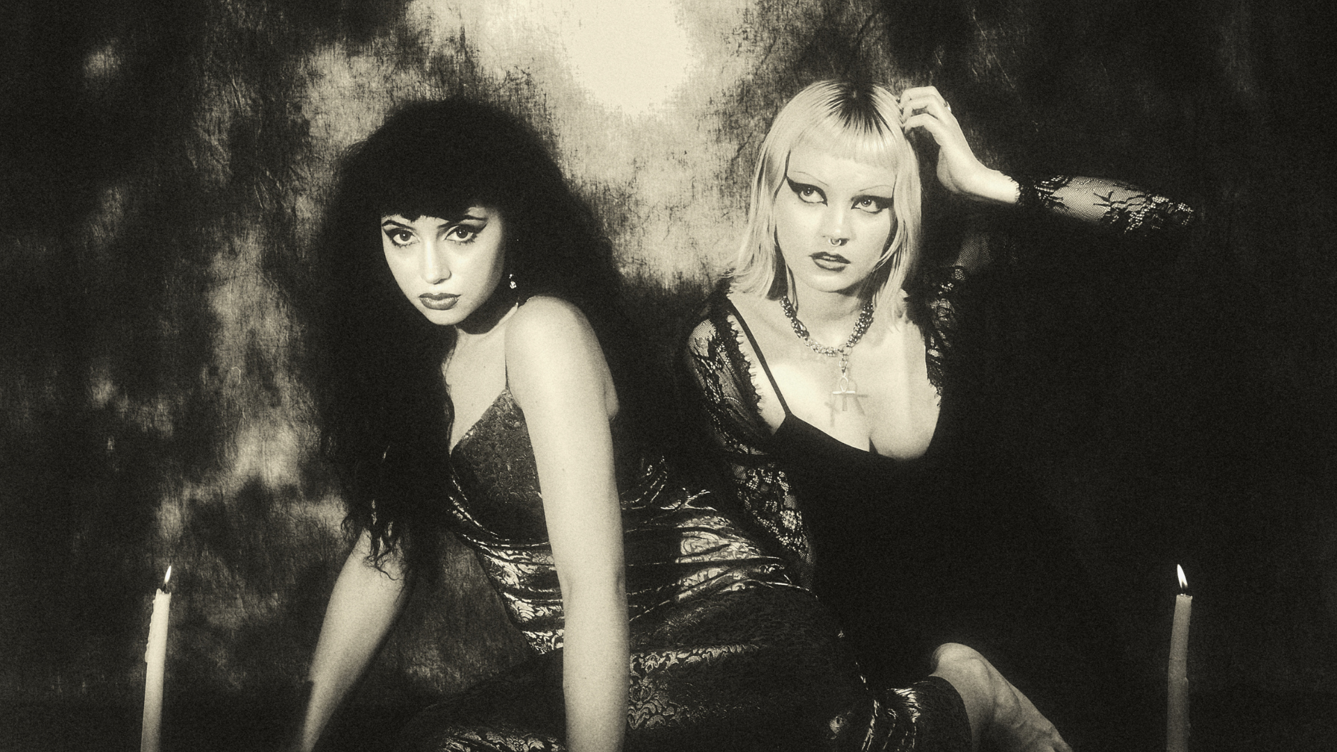 Two women in gothic attire pose dramatically by lit candles, with dark makeup and intense expressions, against a shadowy, textured background in a moody, black-and-white photograph.