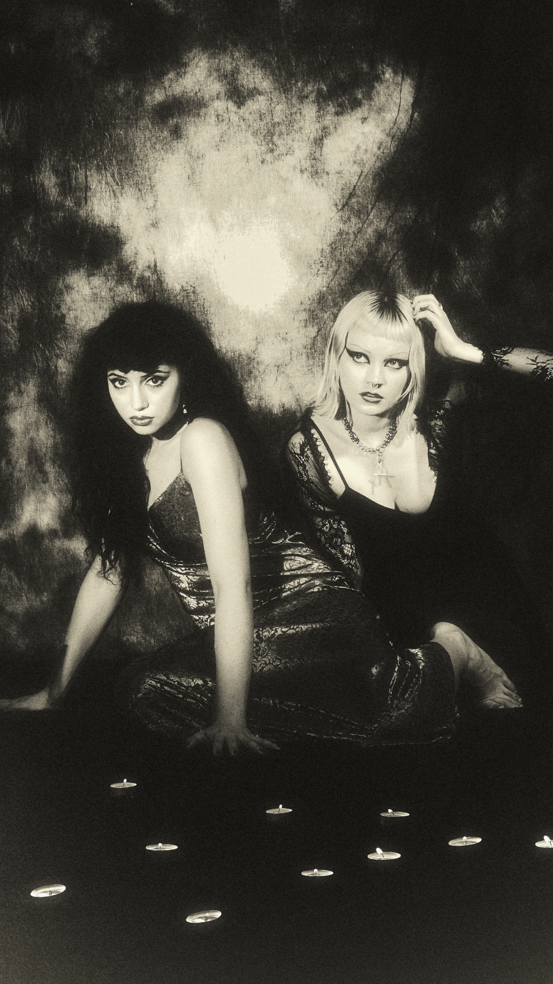 Two women in dark, glamorous outfits sit on the floor in dramatic lighting, surrounded by small lit candles, with a cloudy textured backdrop behind them. The image has a vintage, black-and-white effect.