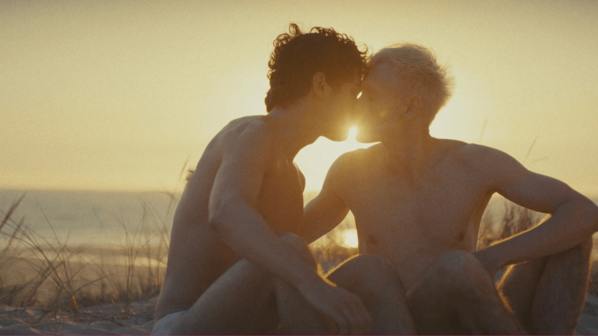 Two shirtless people sit close together on a beach at sunset, gently touching foreheads and noses, with the sunlight softly illuminating them from behind.