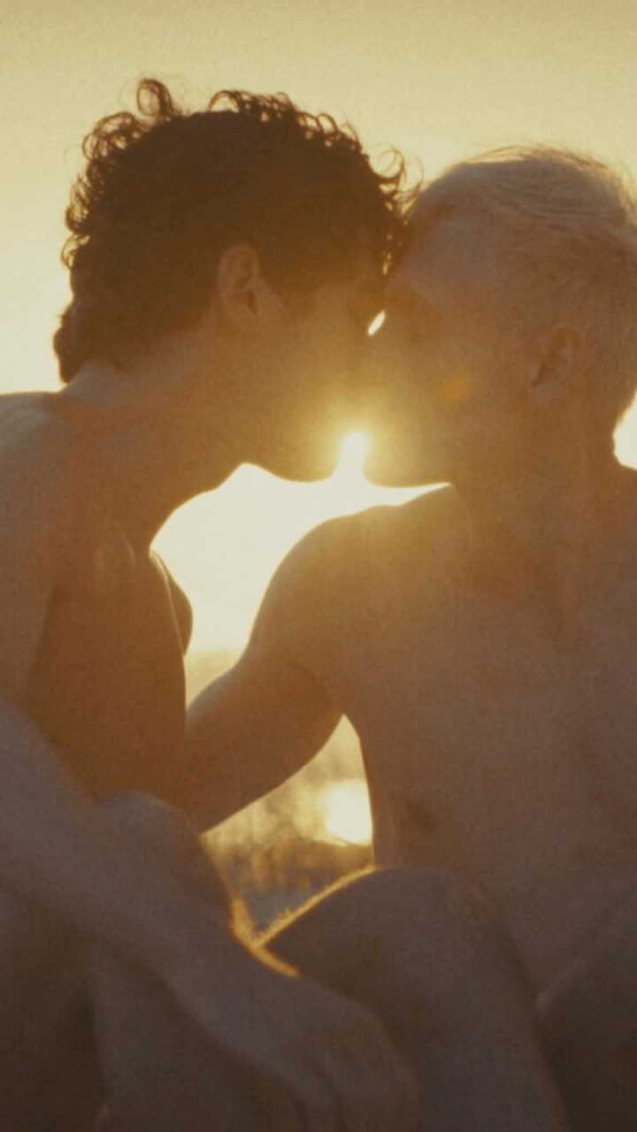 Two shirtless people sit closely together on a beach at sunset, touching foreheads and appearing intimate, with warm sunlight behind them and the ocean in the background.