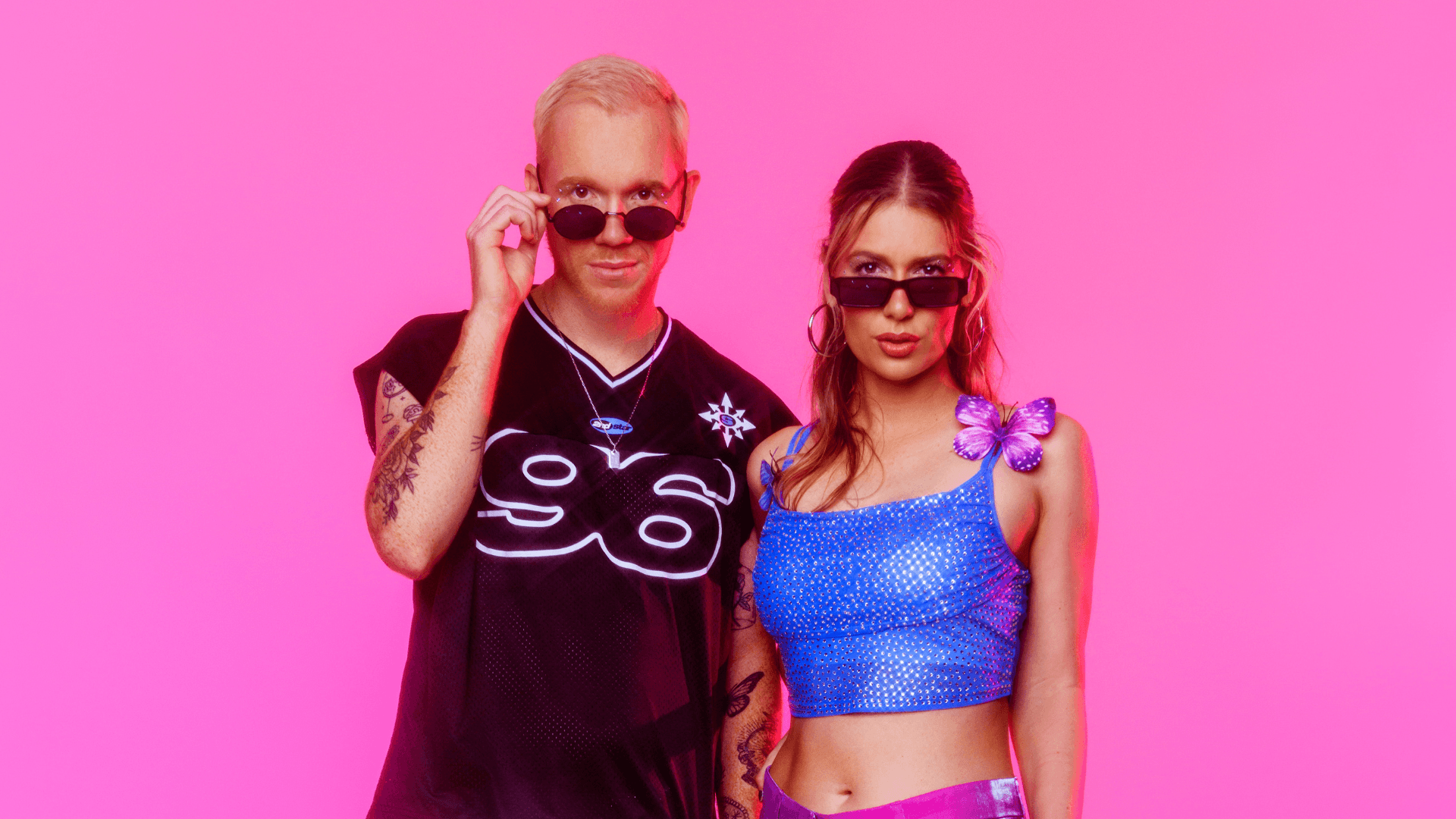 Two people stand side by side against a bright pink background, both wearing sunglasses. The person on the left has short blond hair and a black jersey, while the person on the right has long brown hair and a blue sparkly top with a purple butterfly accessory.