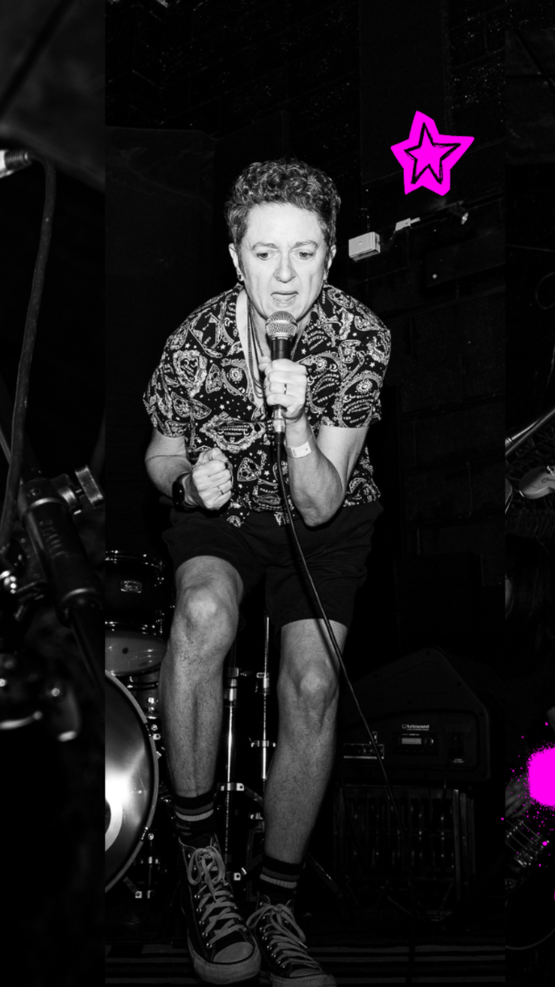 A person in patterned shirt and shorts passionately sings into a microphone on stage; a drum set is visible behind them. Neon pink star and paint accent the black-and-white image.