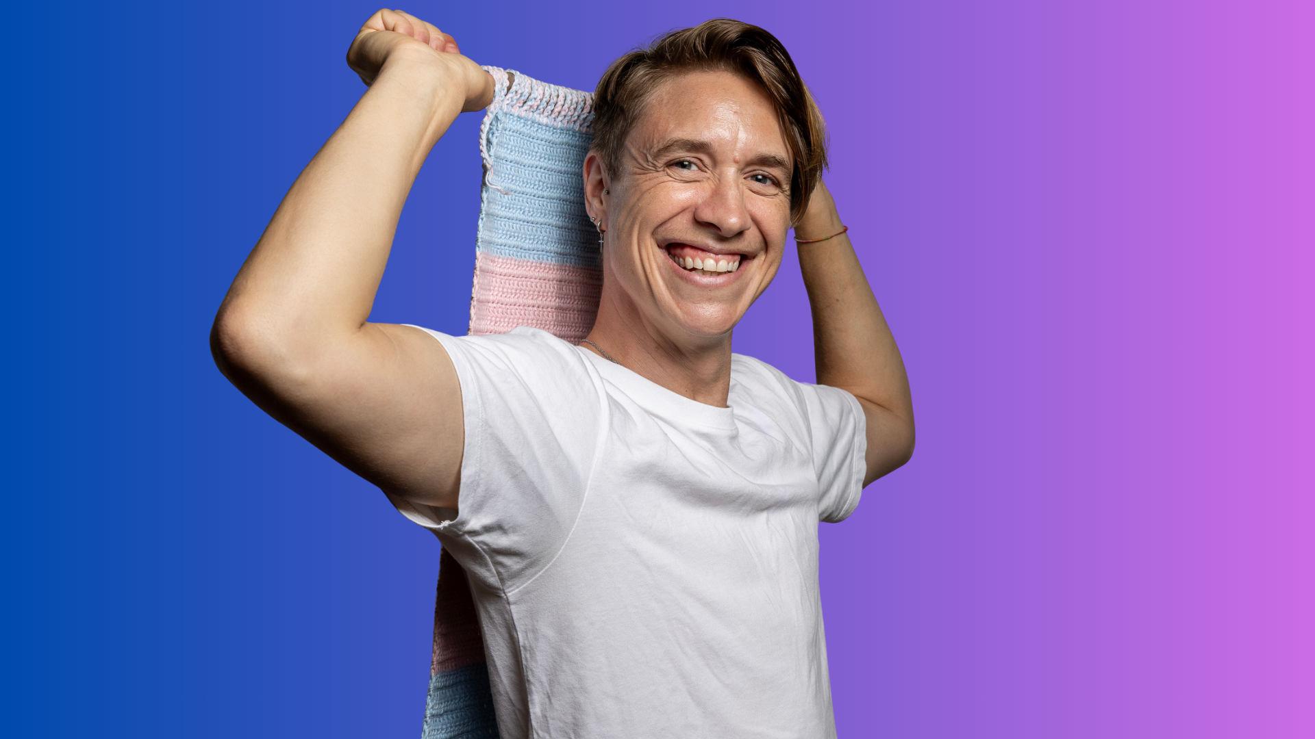 Smiling person in a white shirt holds a blue, pink, and white striped cloth behind their head, standing against a blue to purple gradient background.