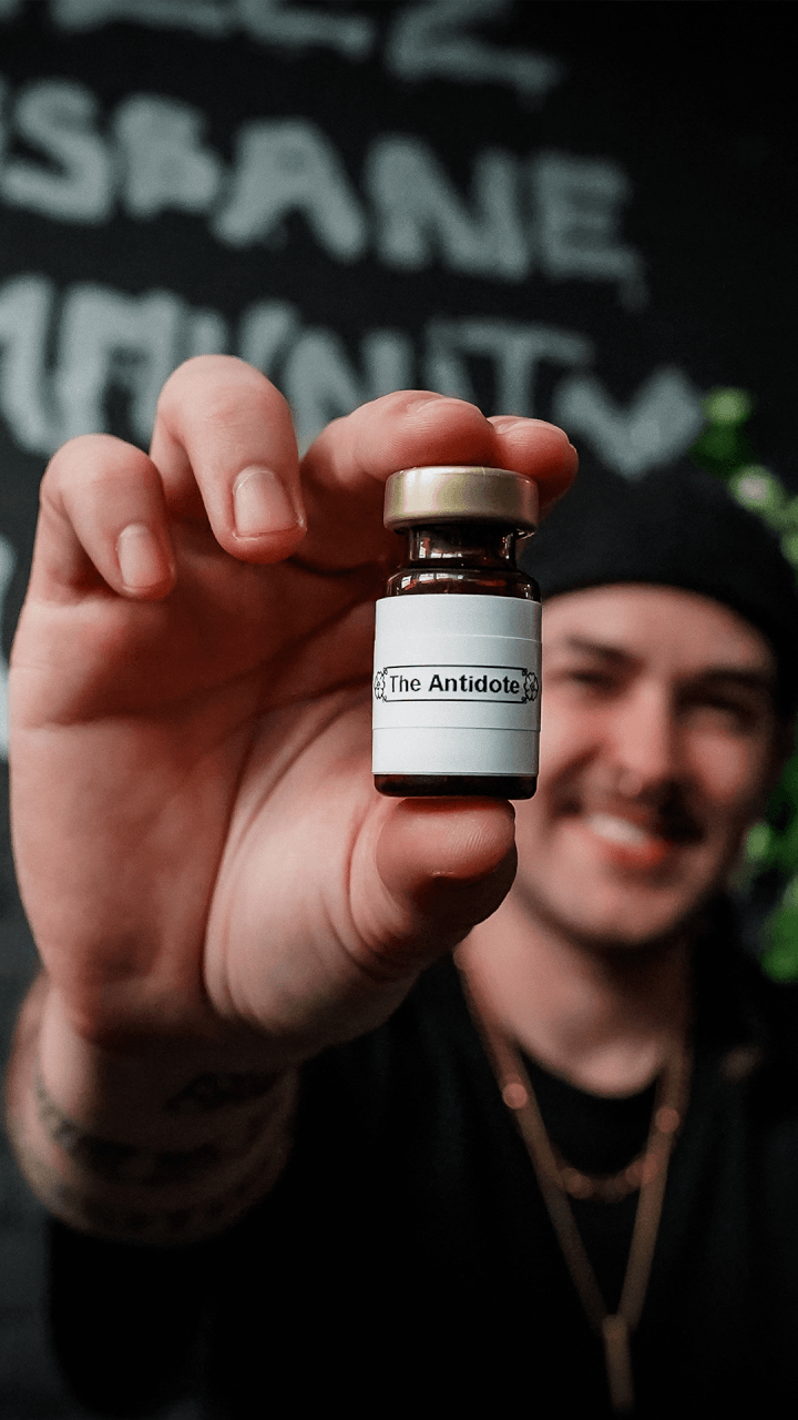 A person holds a small glass vial labeled The Antidote toward the camera, with a blurred smiling person in the background against a dark wall with white text.
