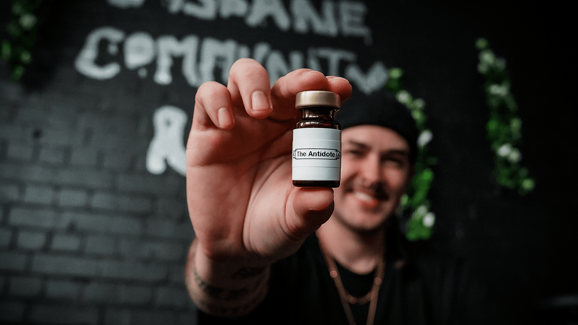 A person smiles while holding a small bottle labeled The Antidote toward the camera, with a blurred black brick wall and some greenery in the background.