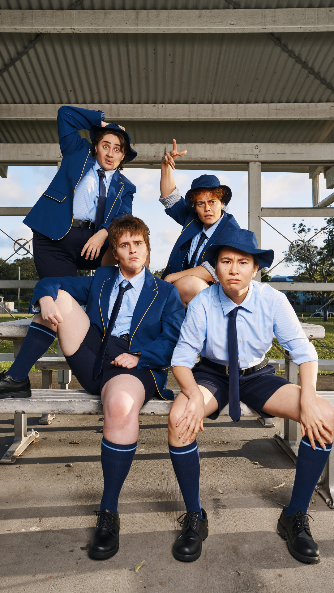 Four people in blue school uniforms and hats pose confidently on bleachers, each displaying playful and expressive stances under a metal roof, with trees and a field visible in the background.