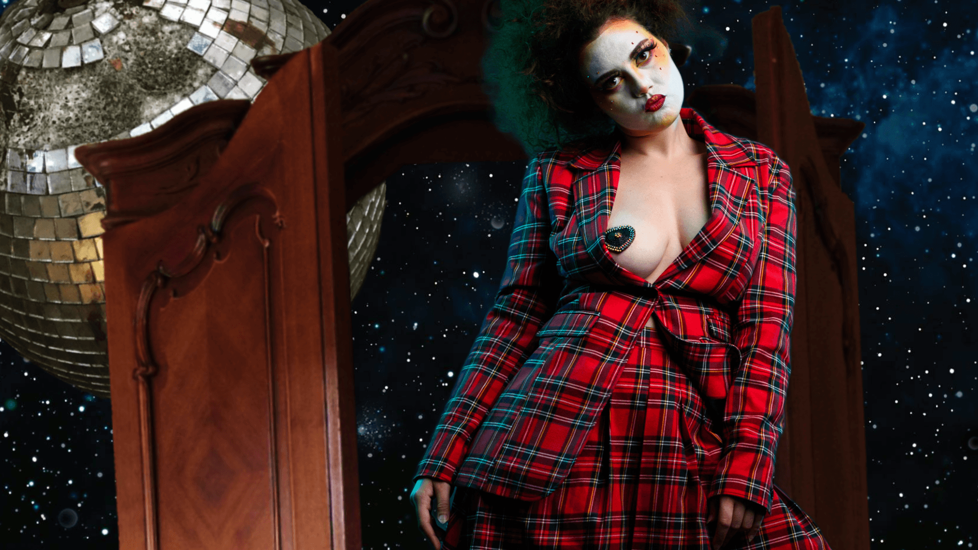 A person in a red plaid suit with dramatic makeup stands before an open wooden wardrobe, revealing a disco ball and starry outer space in the background.