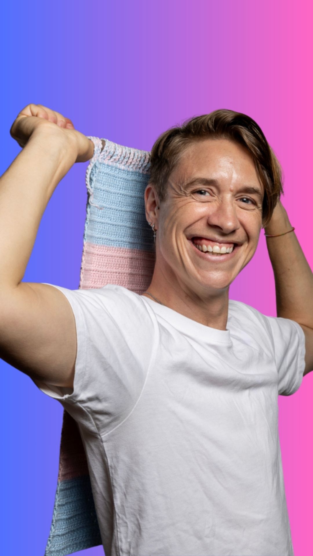 Smiling person in a white shirt holds a pastel pink, blue, and white knit scarf behind their shoulders, standing in front of a vibrant blue-to-pink gradient background.
