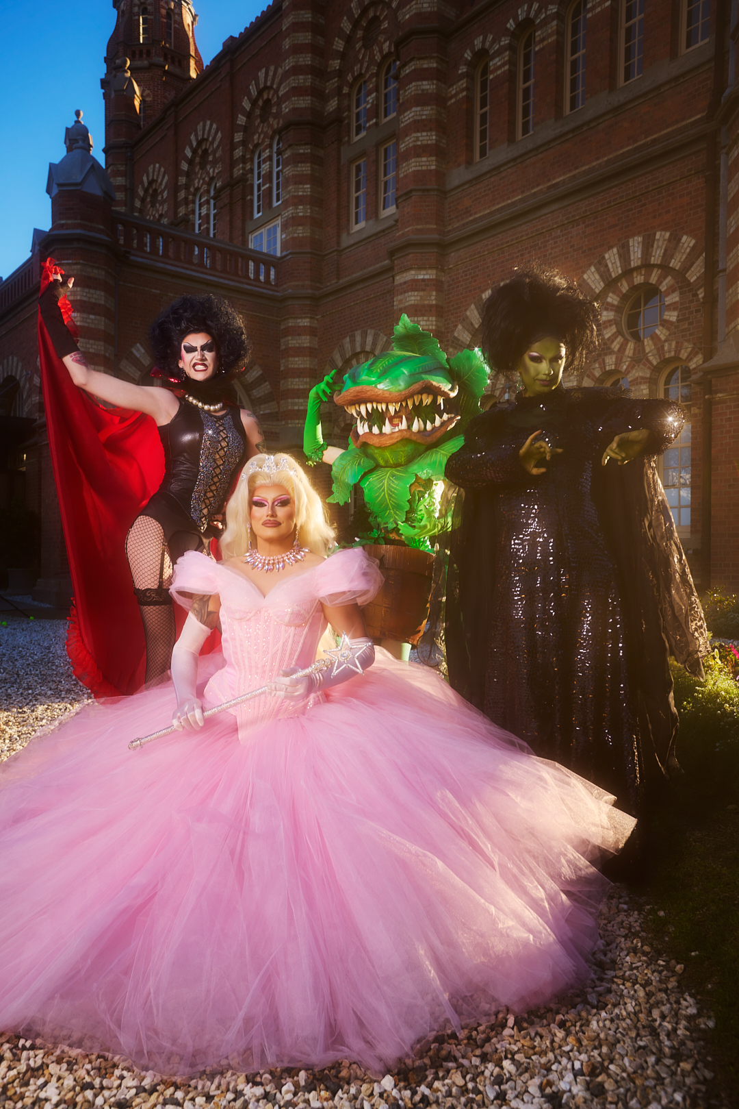 Four people in elaborate costumes stand outside a brick building: one in black with a red cape, one as a green plant, one in a pink gown with a wand, and one in a sparkly black dress with green face paint.