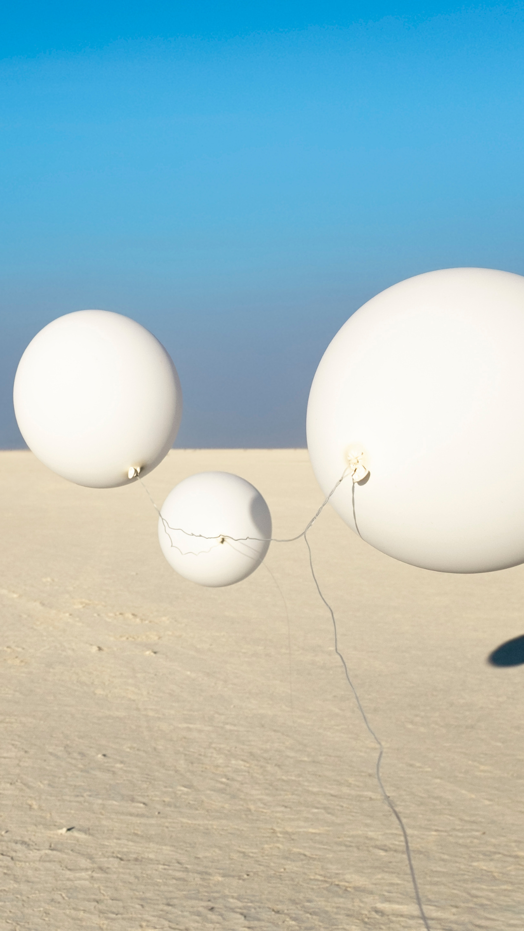 Three large white balloons float above a sandy, flat landscape under a clear blue sky, with thin strings connecting them.