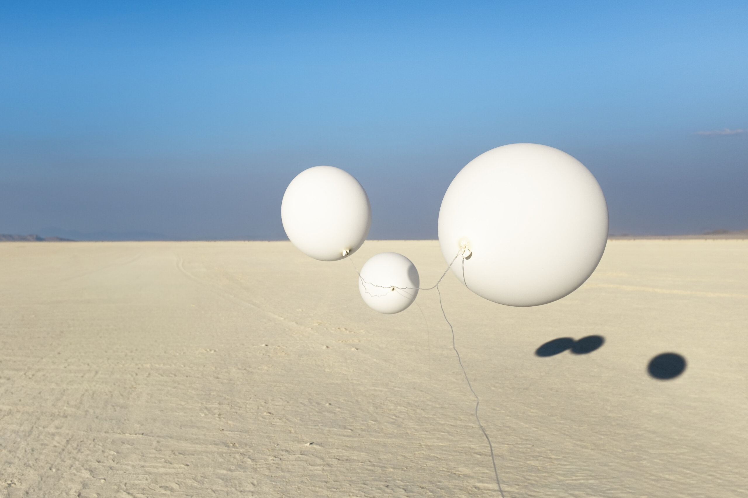 Three large white balloons float above a vast, flat sandy landscape under a clear blue sky. Shadows of the balloons are visible on the ground.