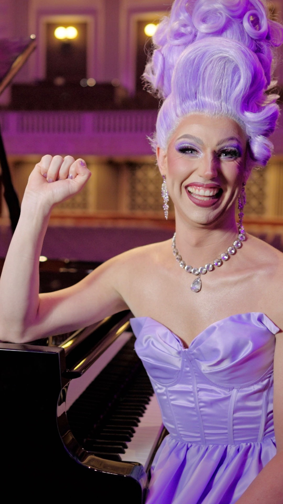 A person with styled lavender hair, dramatic makeup, and a strapless lavender gown smiles while sitting at a piano, wearing sparkling earrings and a necklace.