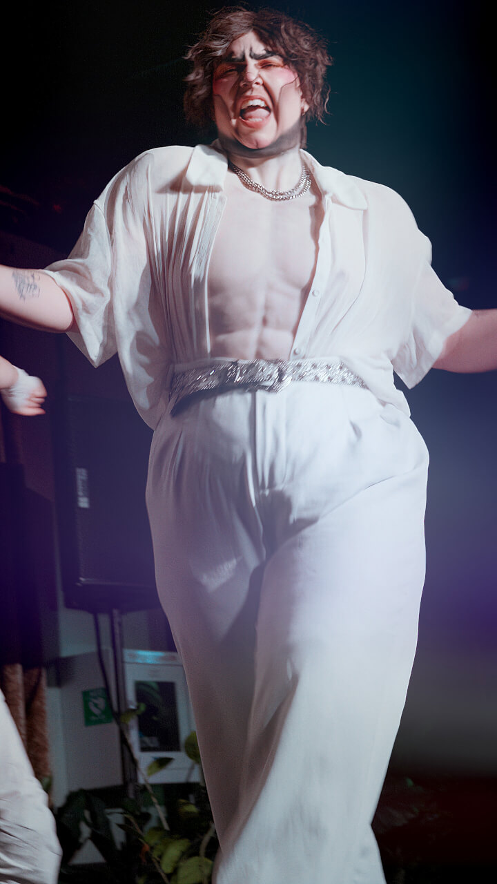 A person in a white, loose outfit and silver belt dances energetically on stage under dramatic lighting, with an expressive face and short, tousled hair.