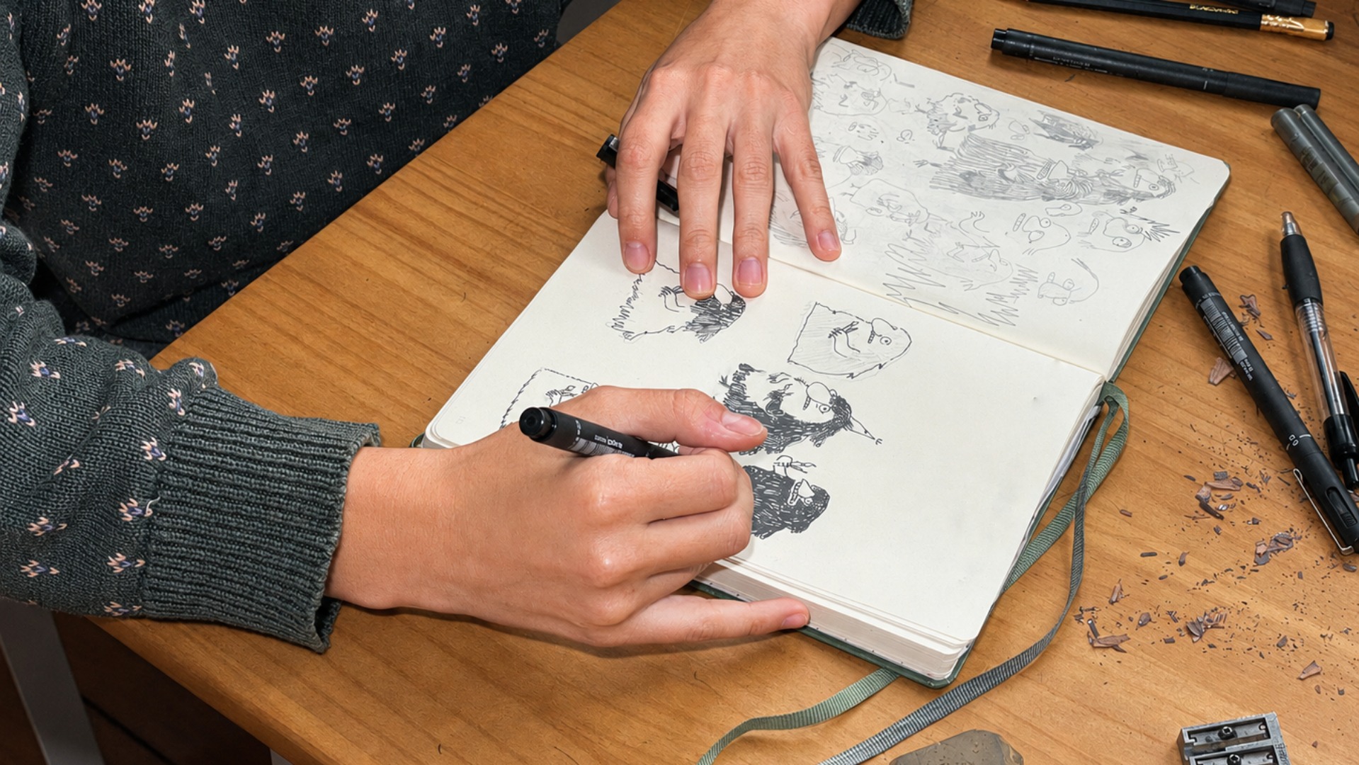 A person sketches cartoonish faces in a notebook using a black pen, surrounded by other pens and drawing tools on a wooden table.