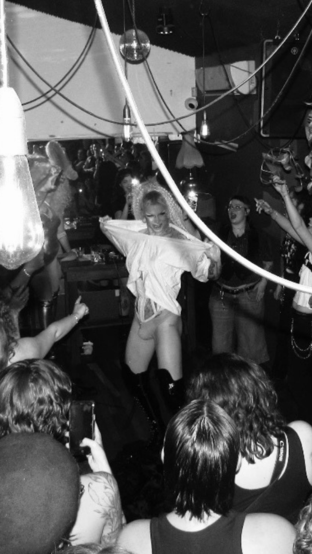 A performer in a white shirt and boots dances in a crowded bar, surrounded by cheering, smiling people. The scene is energetic, with exposed light bulbs and people capturing the moment on their phones. The image is in black and white.
