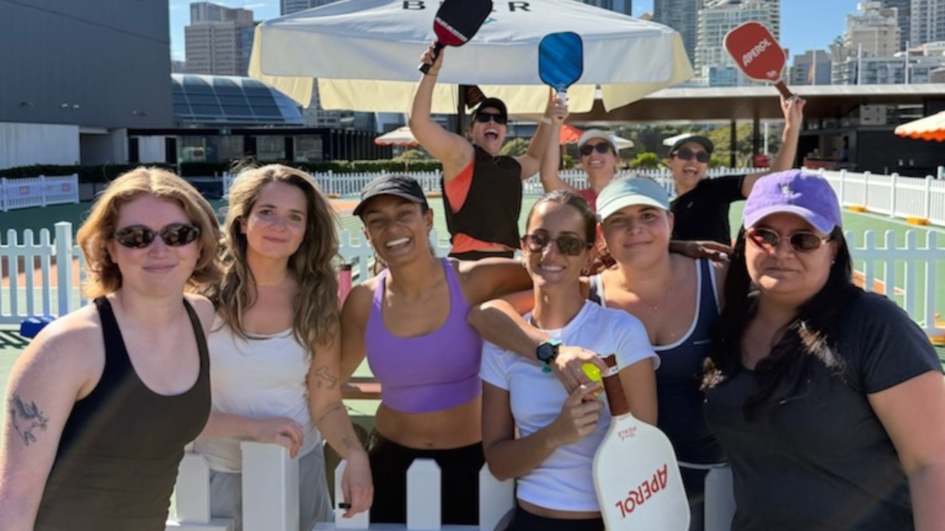 A group of eight women pose and smile outdoors at a pickleball court, some holding paddles, with city buildings in the background on a sunny day.