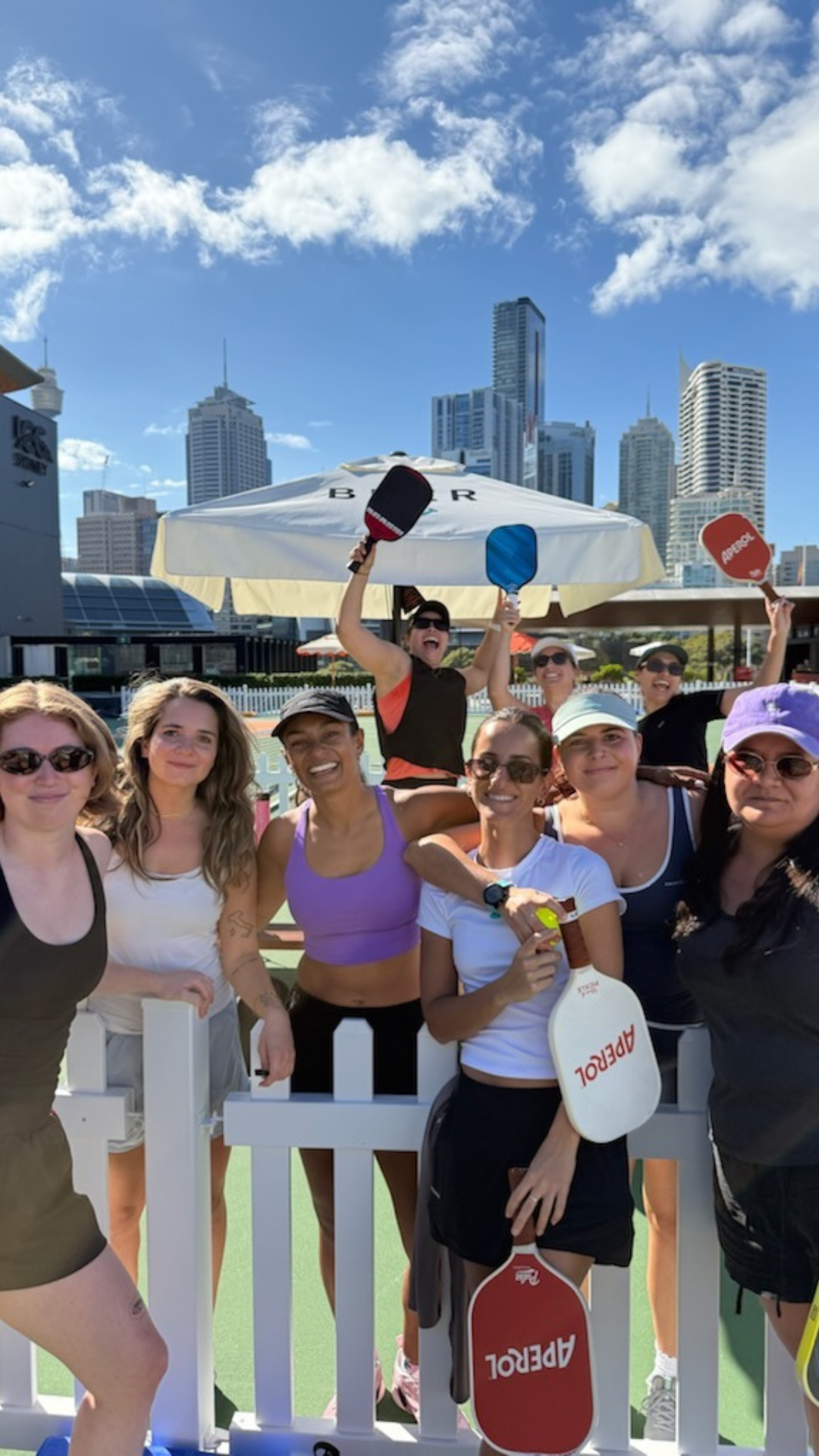 A group of eight smiling people holding pickleball paddles pose in front of a white fence, with city skyscrapers, blue sky, and a large outdoor umbrella in the background.