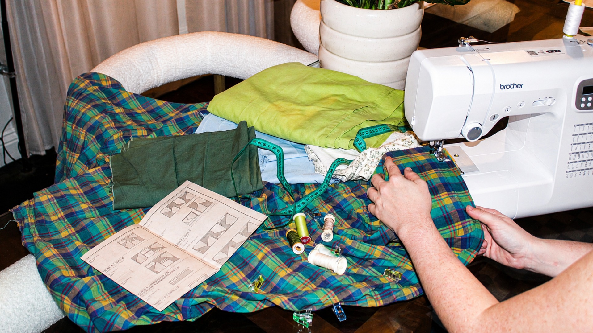 A person uses a sewing machine with green and blue plaid fabric, spools of thread, measuring tape, folded cloth, and sewing instructions laid out nearby.