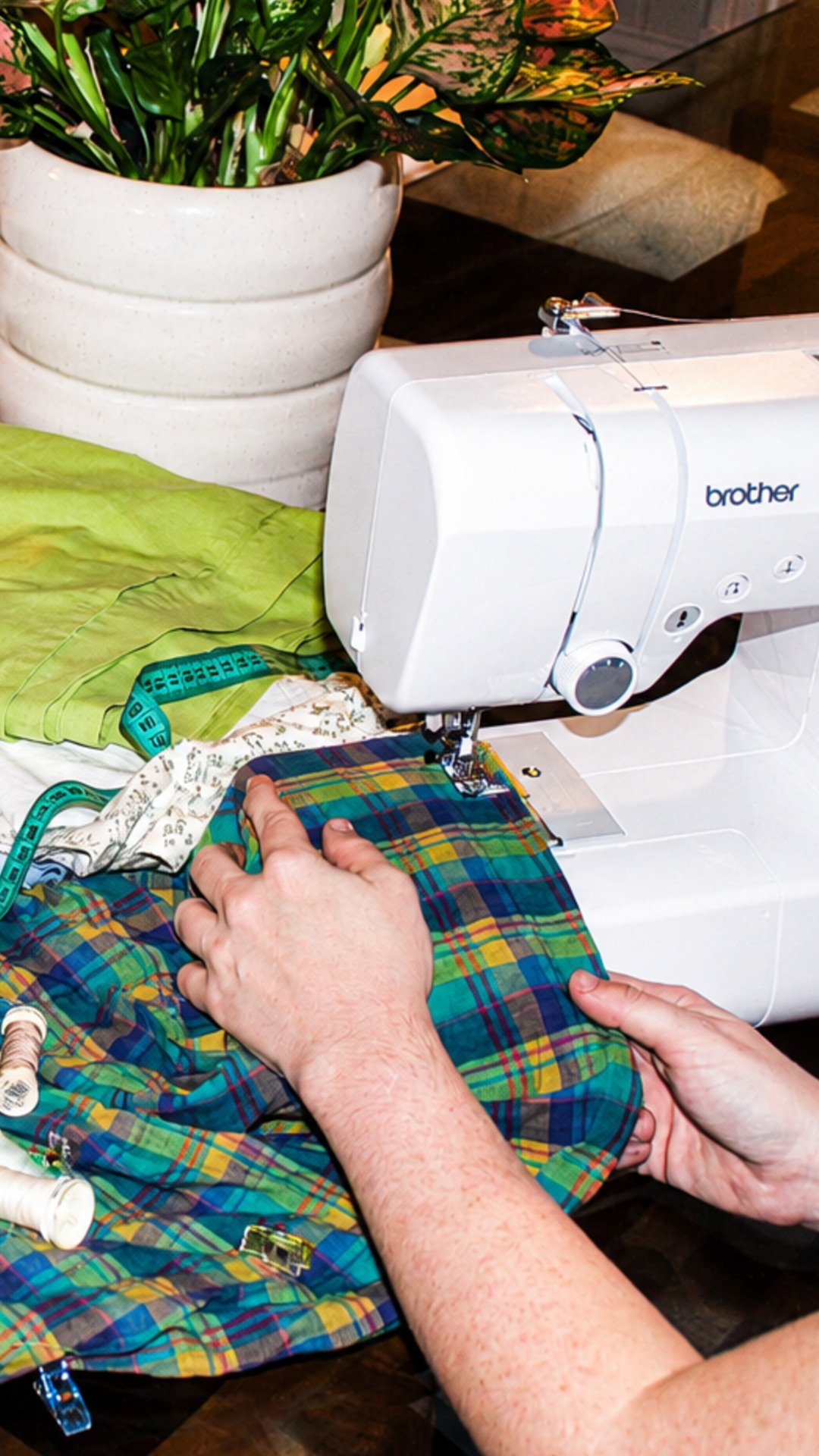 A person sews colorful plaid fabric with a white sewing machine. A green measuring tape, threads, and fabric pieces are nearby.